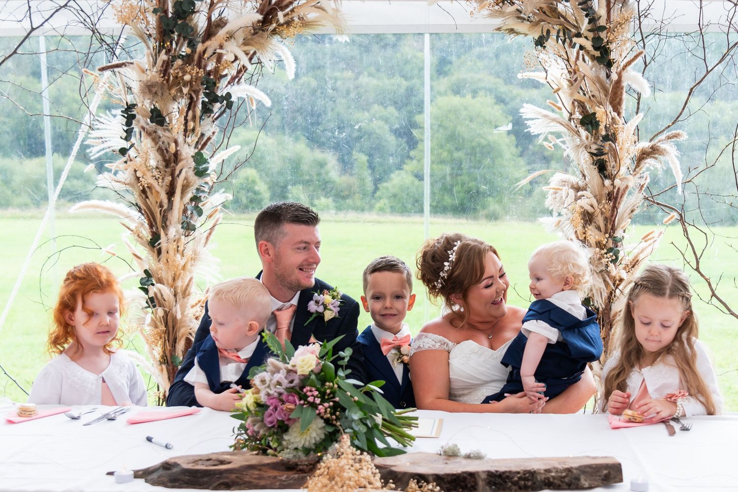 Wedding guests and party members arranged at long table with rustic floral decorations.