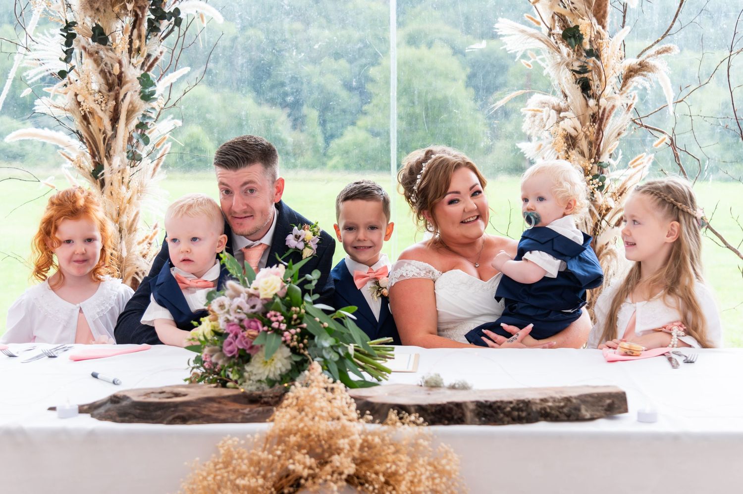 Wedding party group photo with children gathered around a rustic wooden table outdoors.