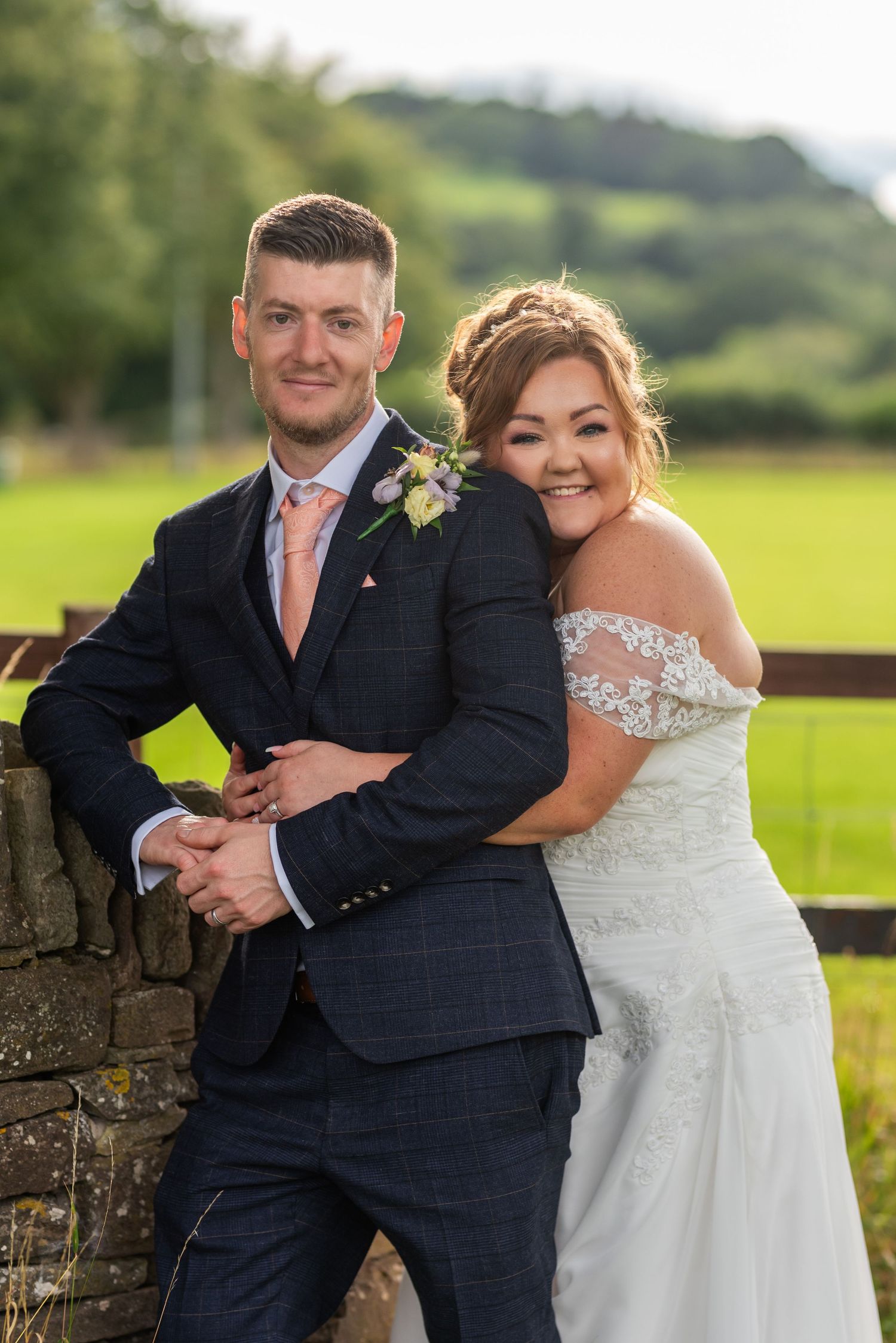 Wedding couple poses playfully against rustic stone wall in countryside setting.
