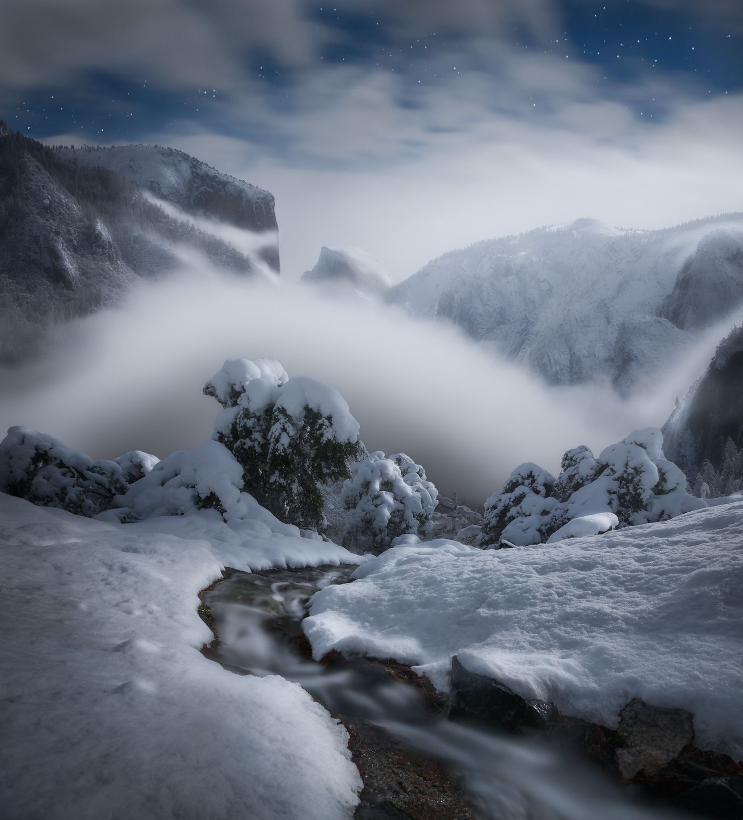 Dramatic winter mountain landscape with snow-covered peaks and misty clouds swirling around a small stream in moody lighting.
