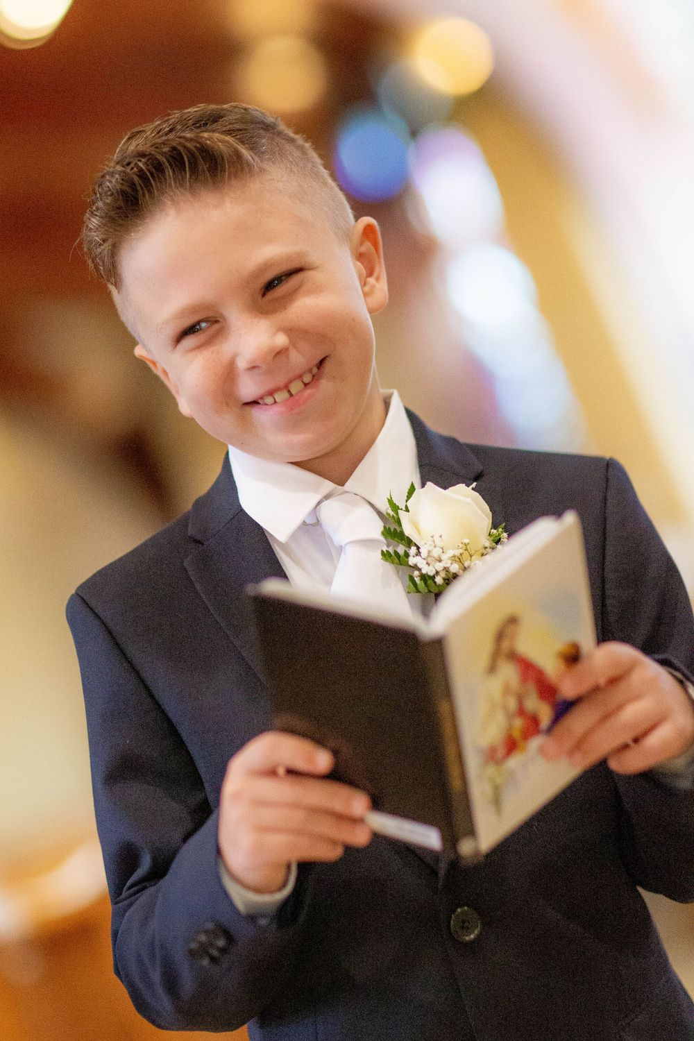A young boy in a dark suit and boutonniere smiles while holding a prayer book in church.