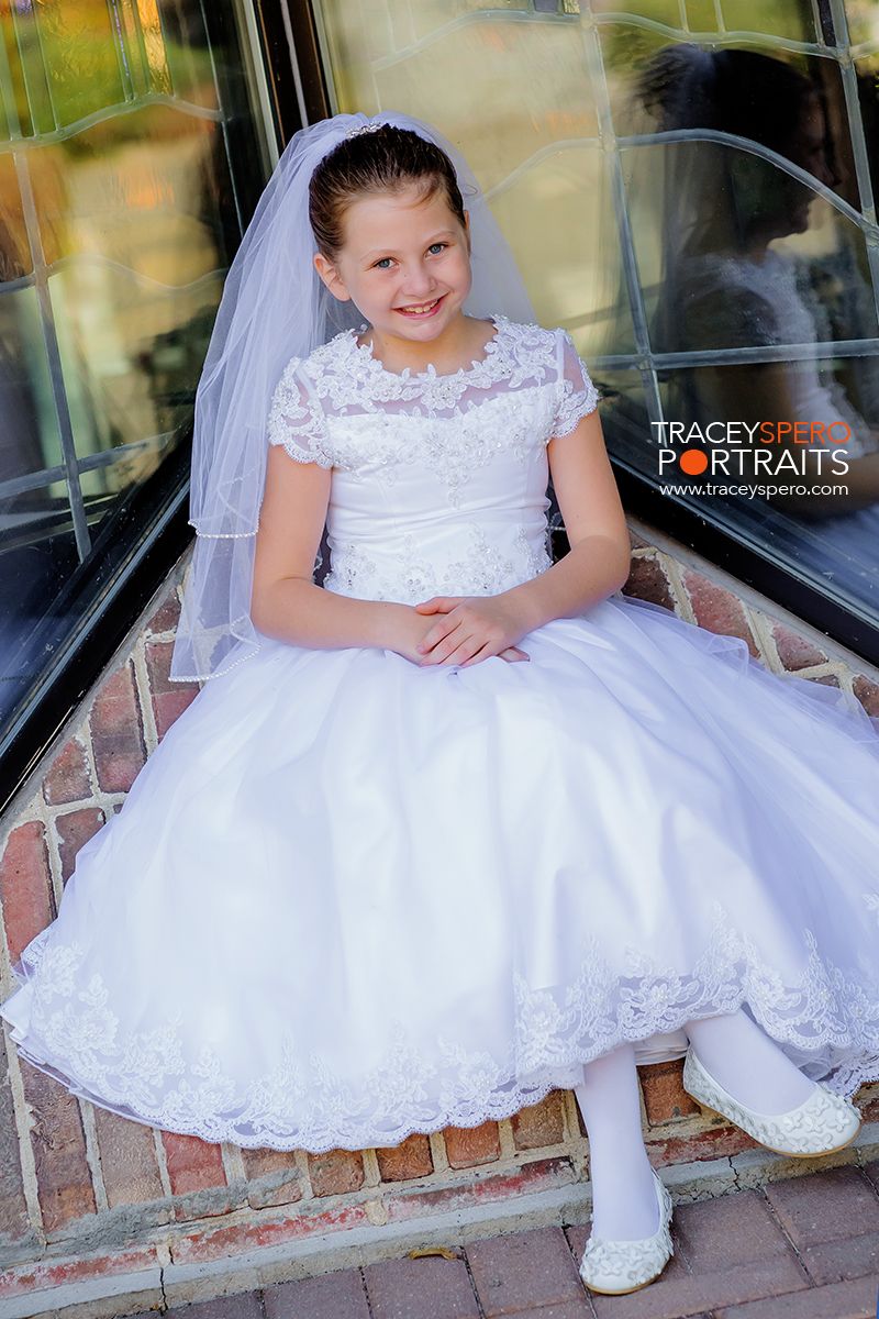 Child in white lace First Communion dress and veil sits on brick ledge.