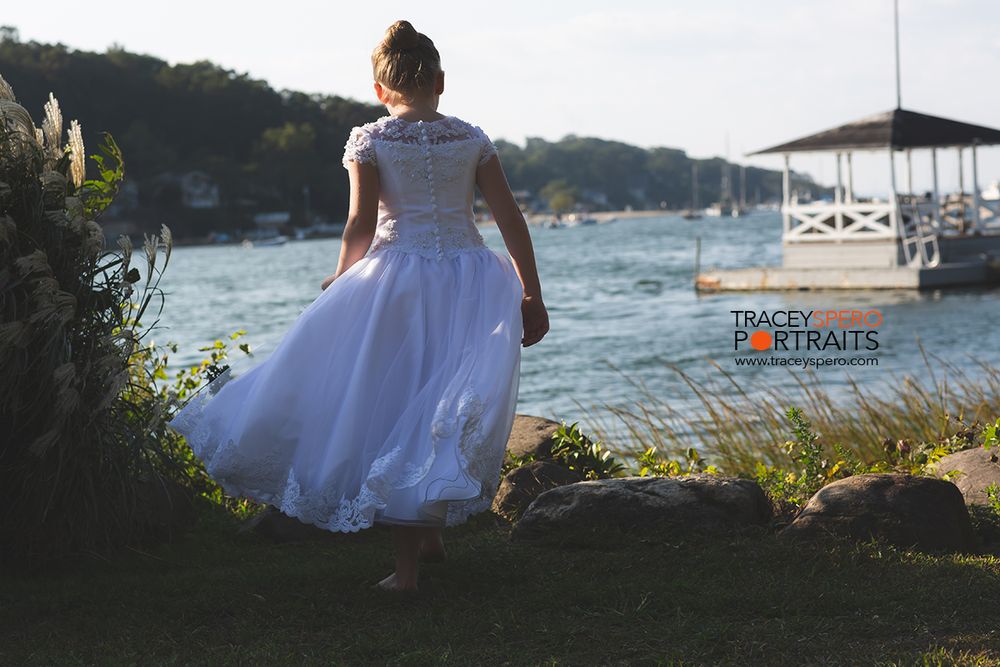 Person in white dress walks along waterfront path with boats in background.