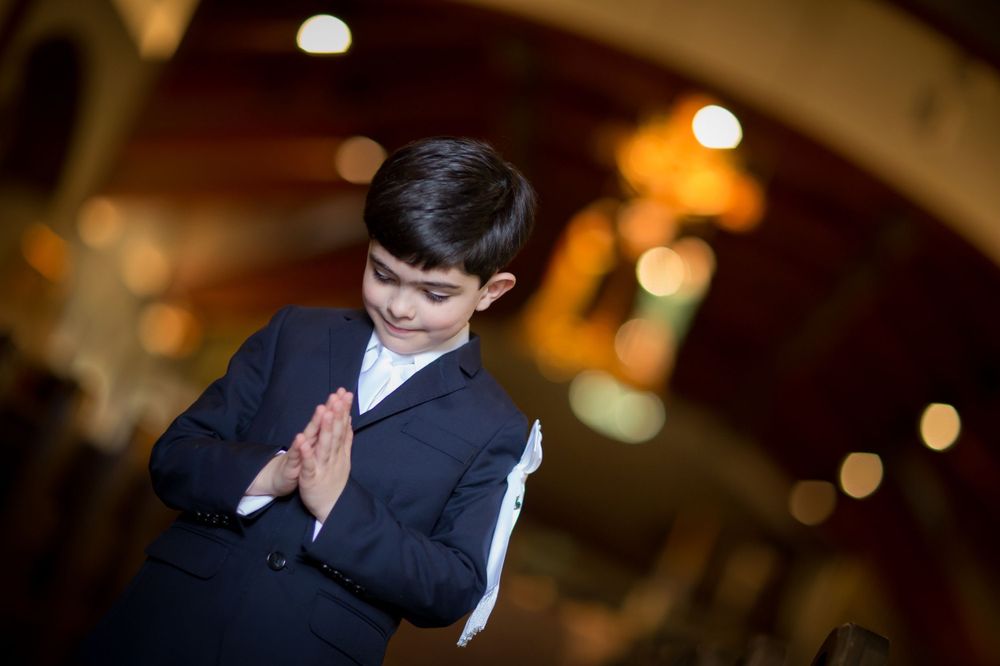 A child in a navy suit prays in an elegant venue with bokeh lights in the background.