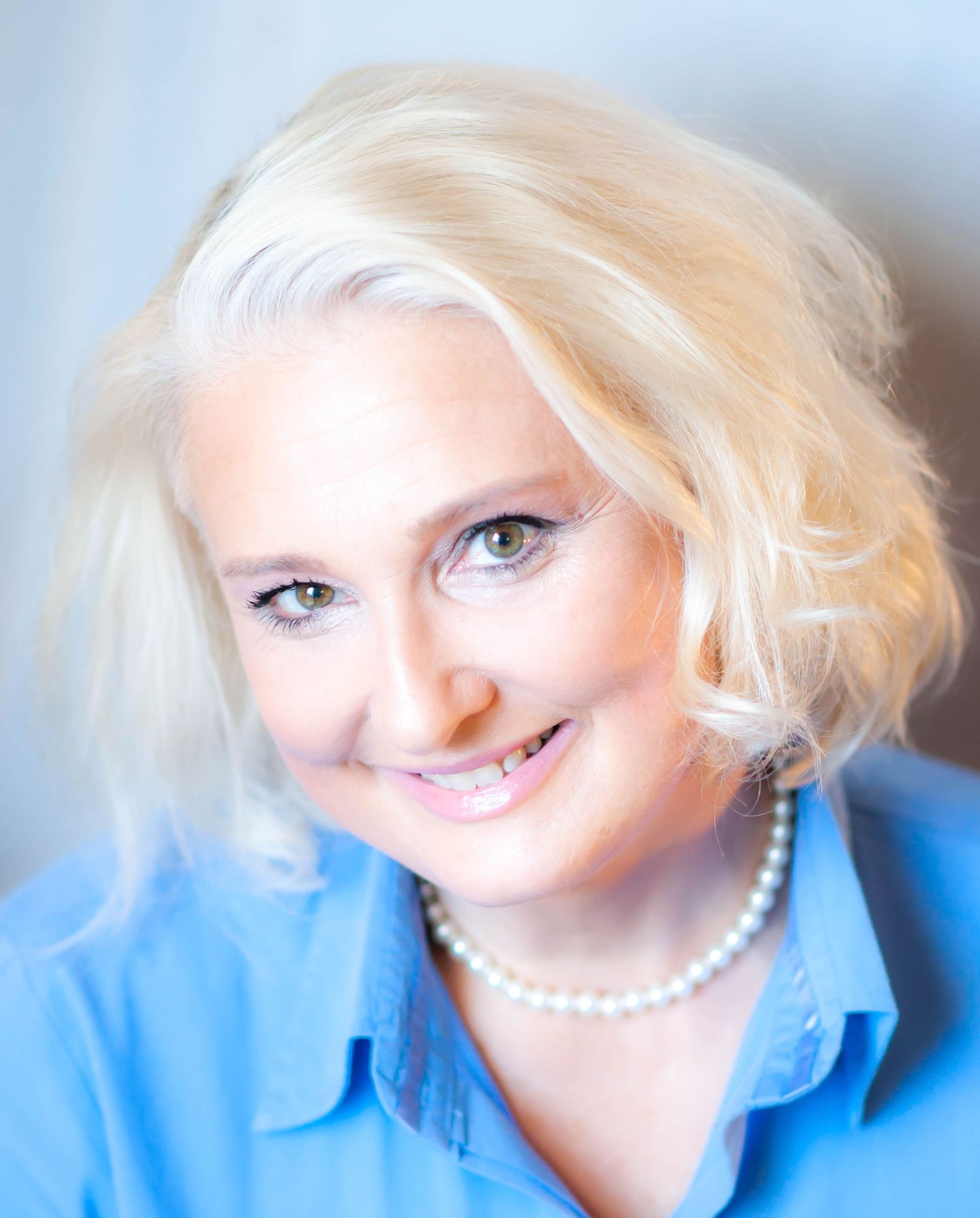Professional headshot of a smiling middle-aged woman with short blonde hair wearing a blue shirt and pearl necklace.