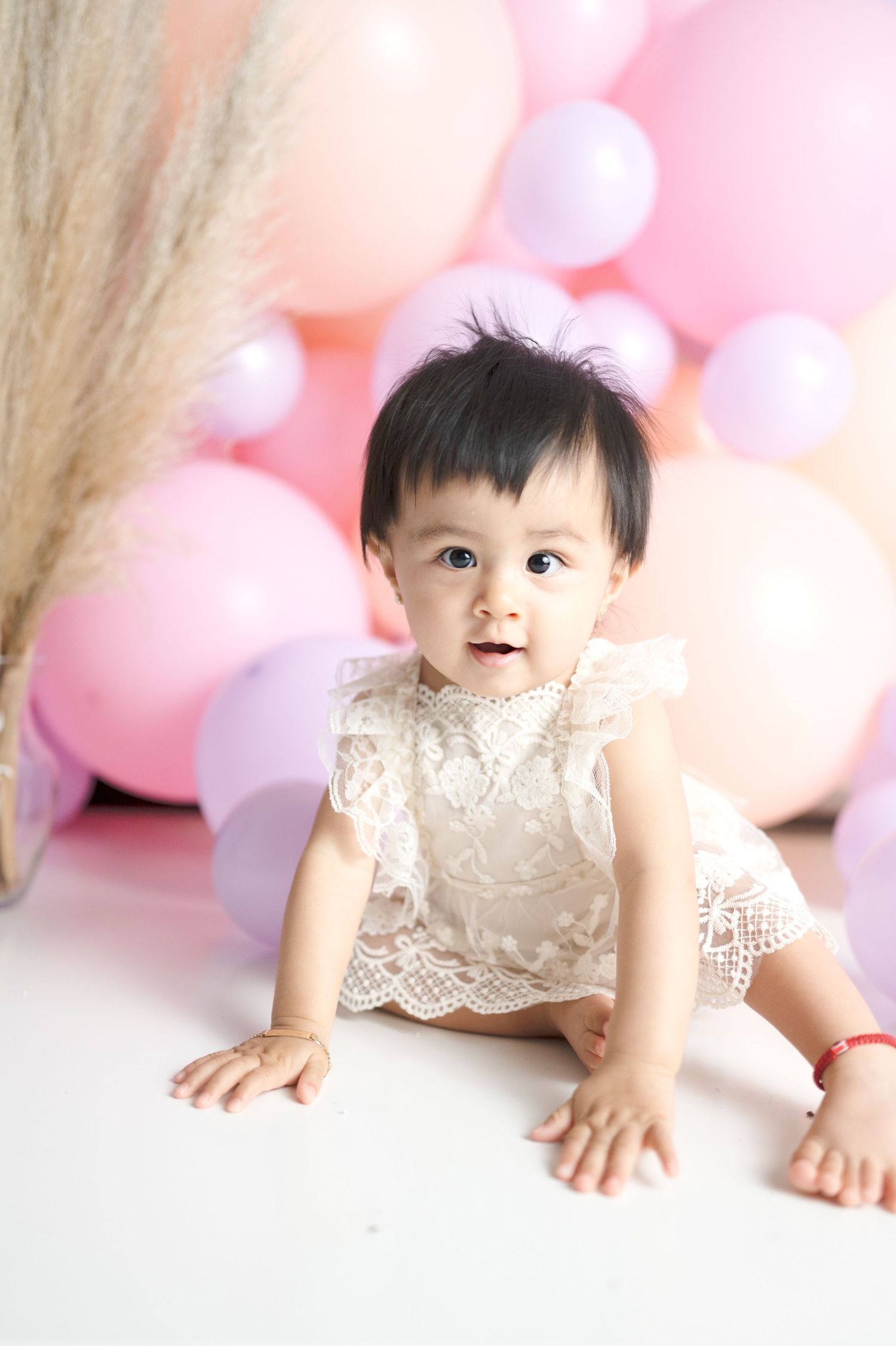 Baby in lace outfit sits against pink balloon backdrop during studio photoshoot.