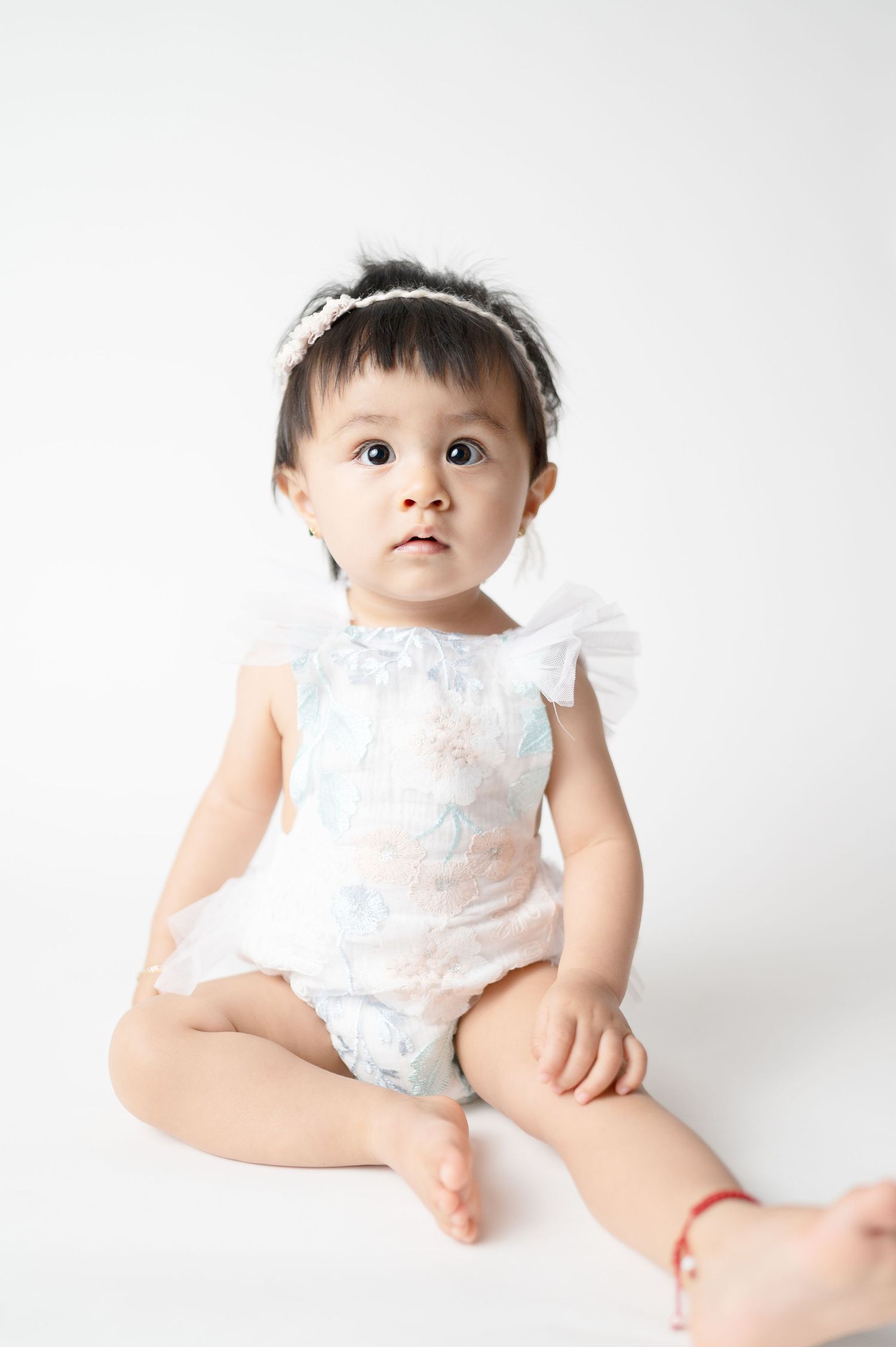 Baby in white outfit sits on plain backdrop during minimalist studio portrait.