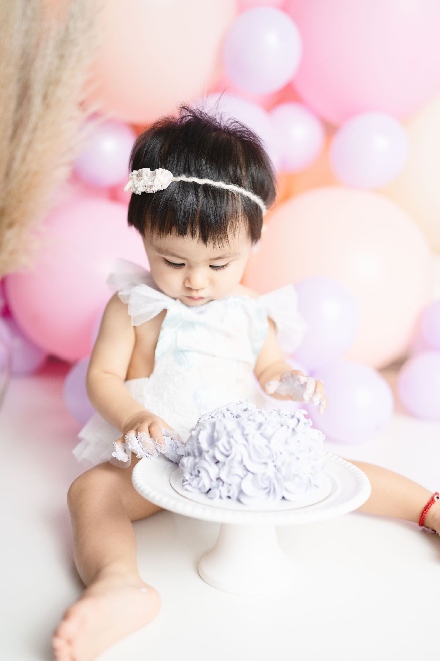 Adorable cake smash setup with pink balloons and white dress against pastel backdrop.