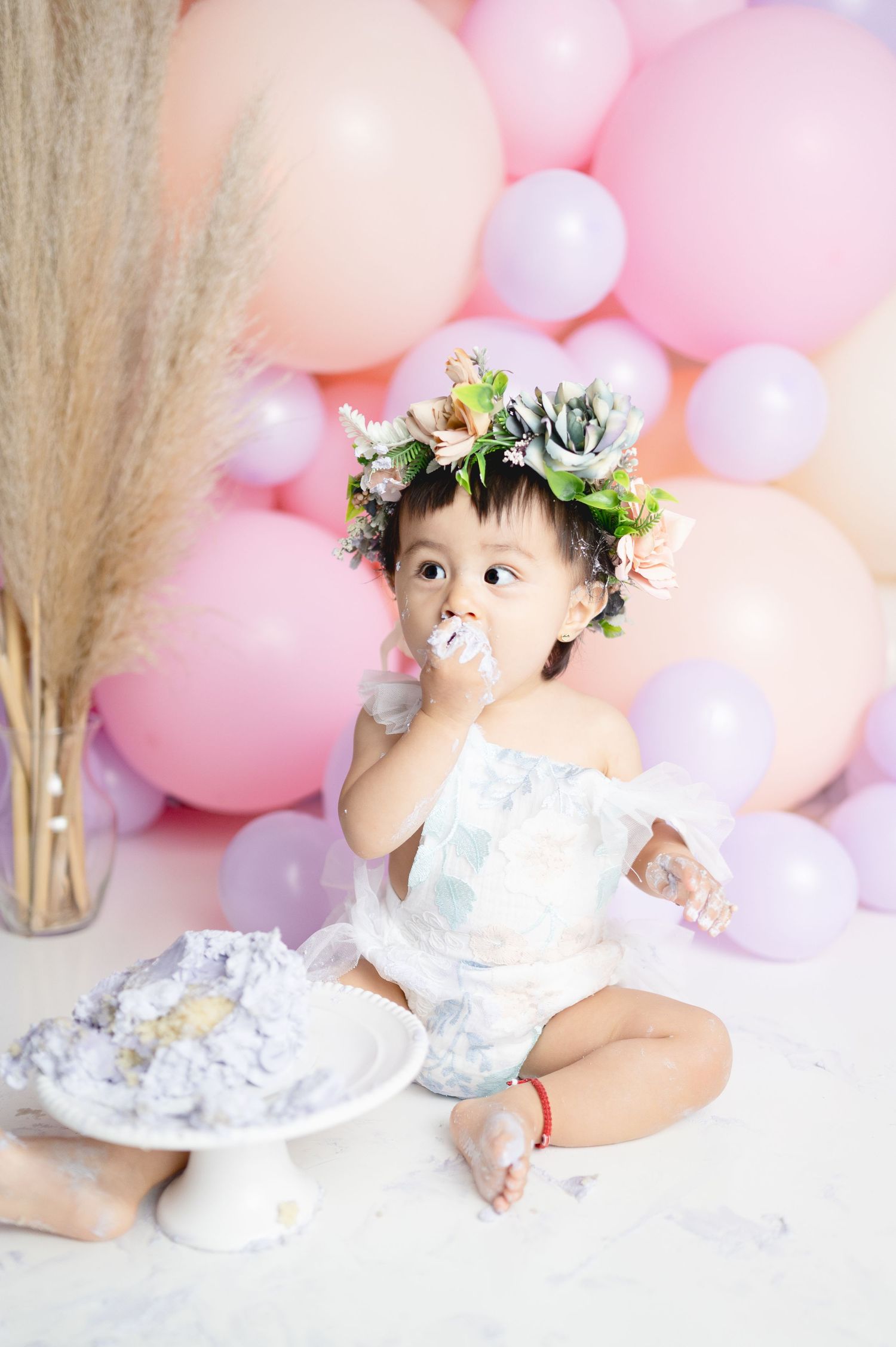 Baby in white dress and floral crown enjoying cake during first birthday photoshoot.