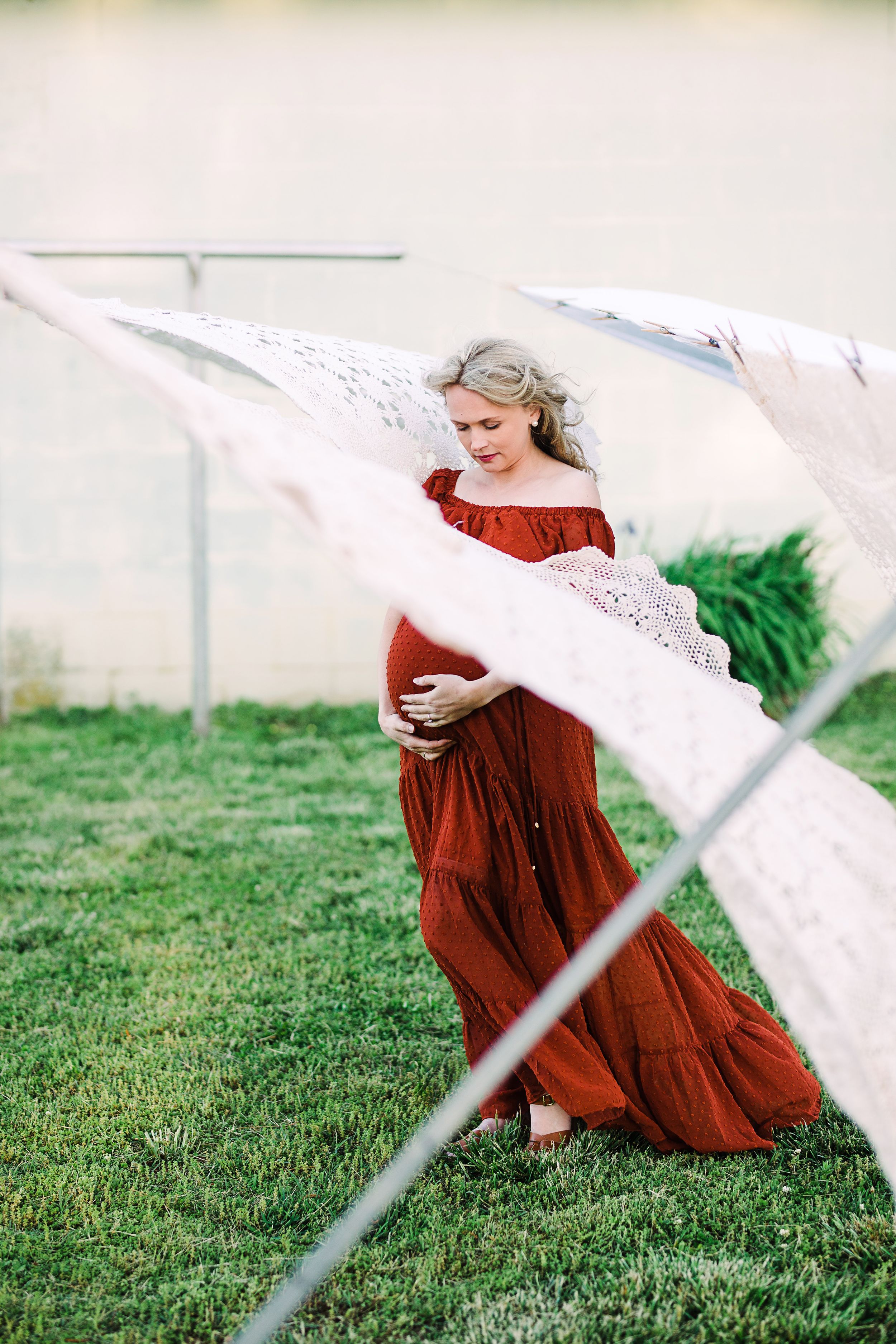 Pregnant woman in rust red off-shoulder gown holding belly near hanging white lace fabric outdoors.