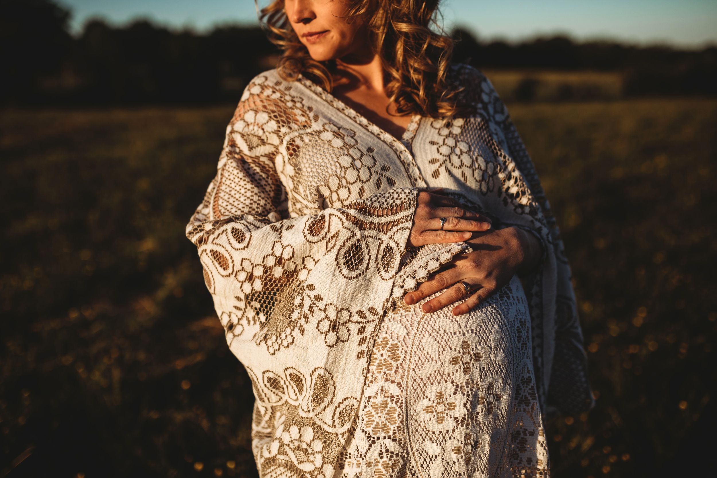 Close-up of pregnant woman in ornate lace robe cradling her baby bump in a golden field.