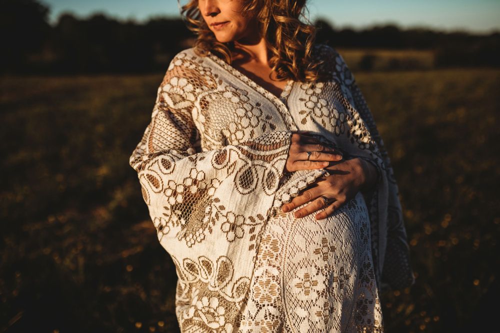 Close-up of pregnant woman in ornate lace robe cradling her baby bump in a golden field.