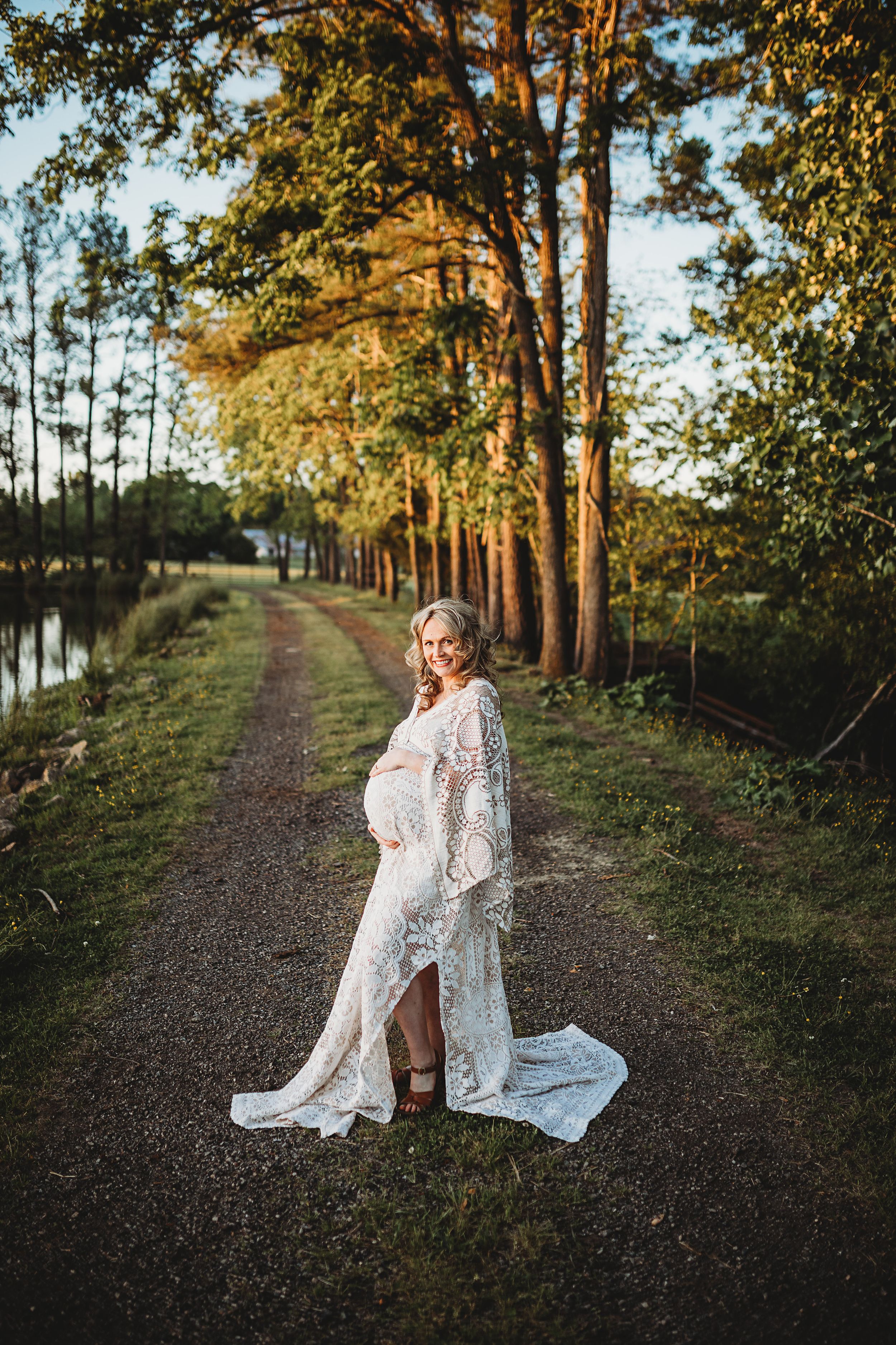 Pregnant woman in white lace gown smiling on tree-lined gravel path at sunset.