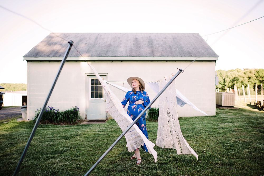 Pregnant woman in blue floral dress and hat hanging laundry outside cottage.