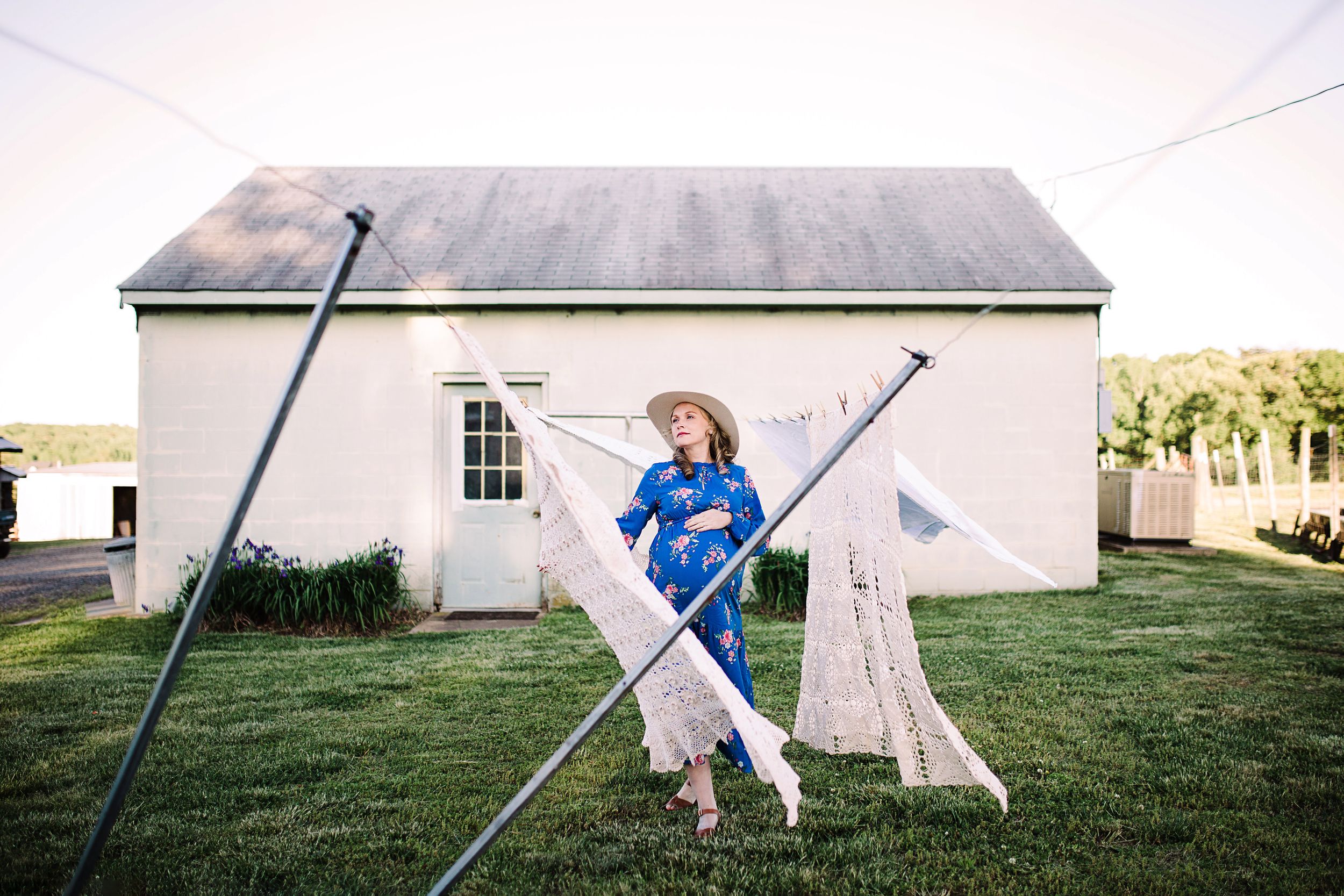 Pregnant woman in blue floral dress and hat hanging laundry outside cottage.