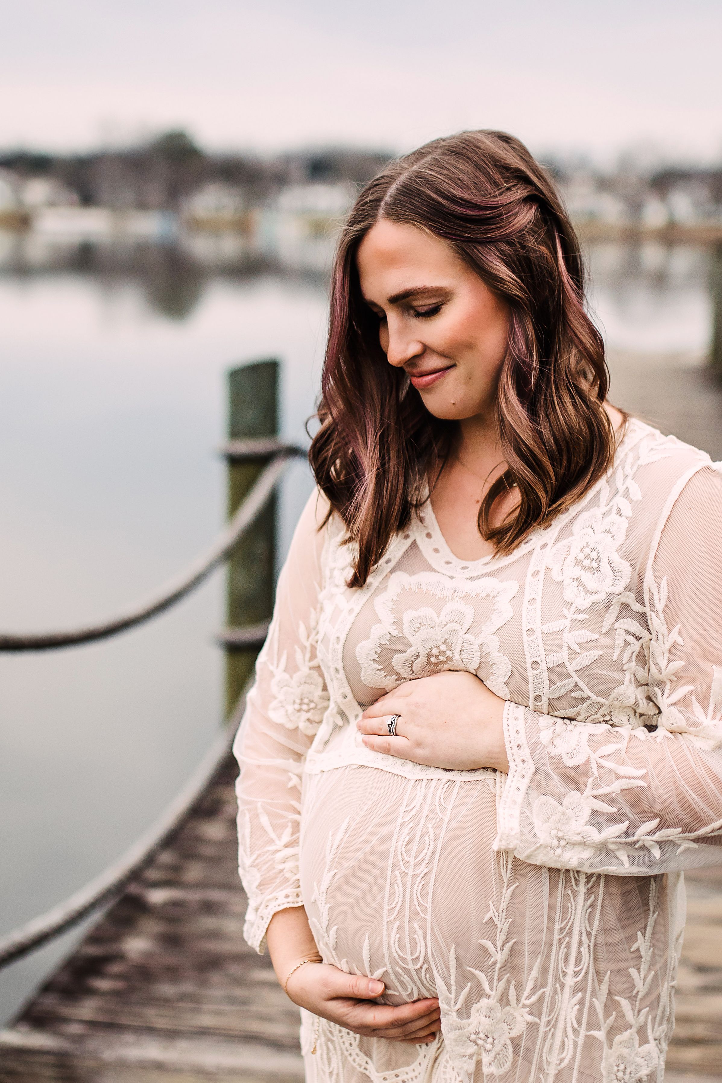 Elegant pregnant woman in lace dress holding baby bump on waterfront dock at dusk.