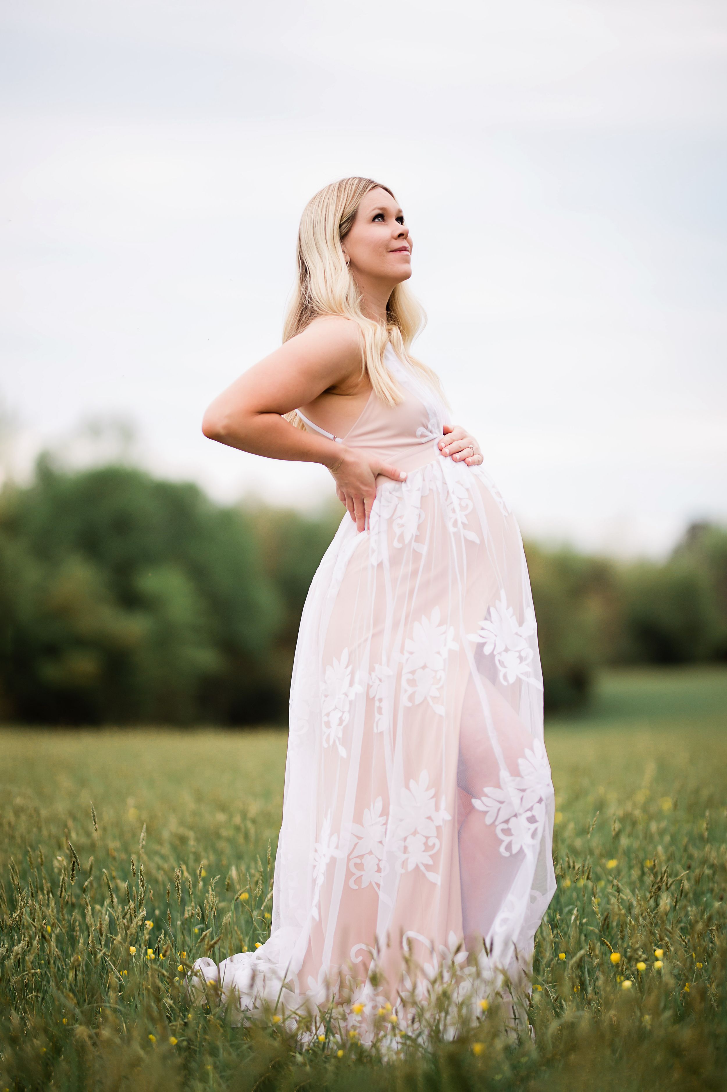 Blonde pregnant woman in sheer pink lace maternity gown standing in green meadow field.