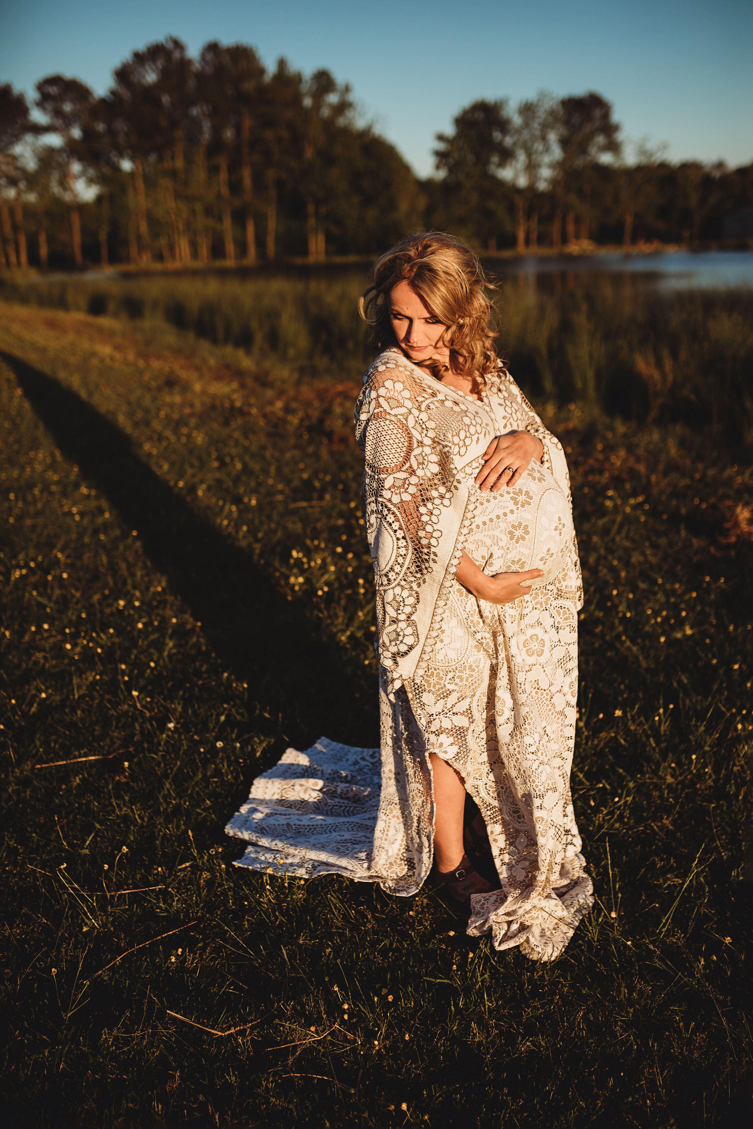 Pregnant woman wrapped in lace shawl standing in golden field near lake.
