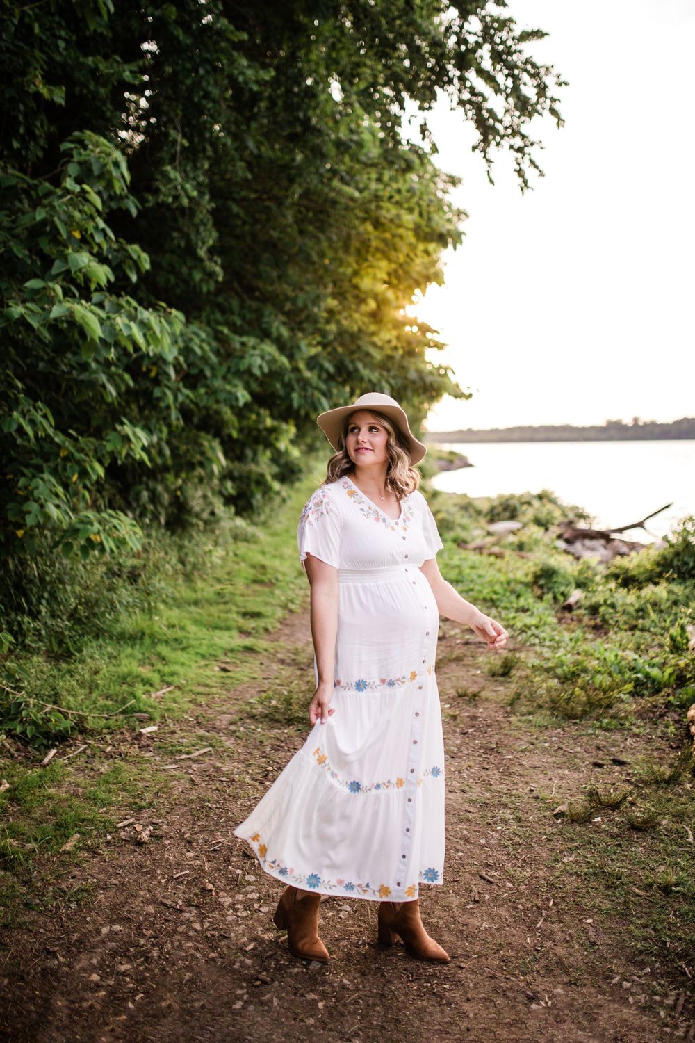 Pregnant woman in white embroidered maxi dress and straw hat walking along a lakeside trail.