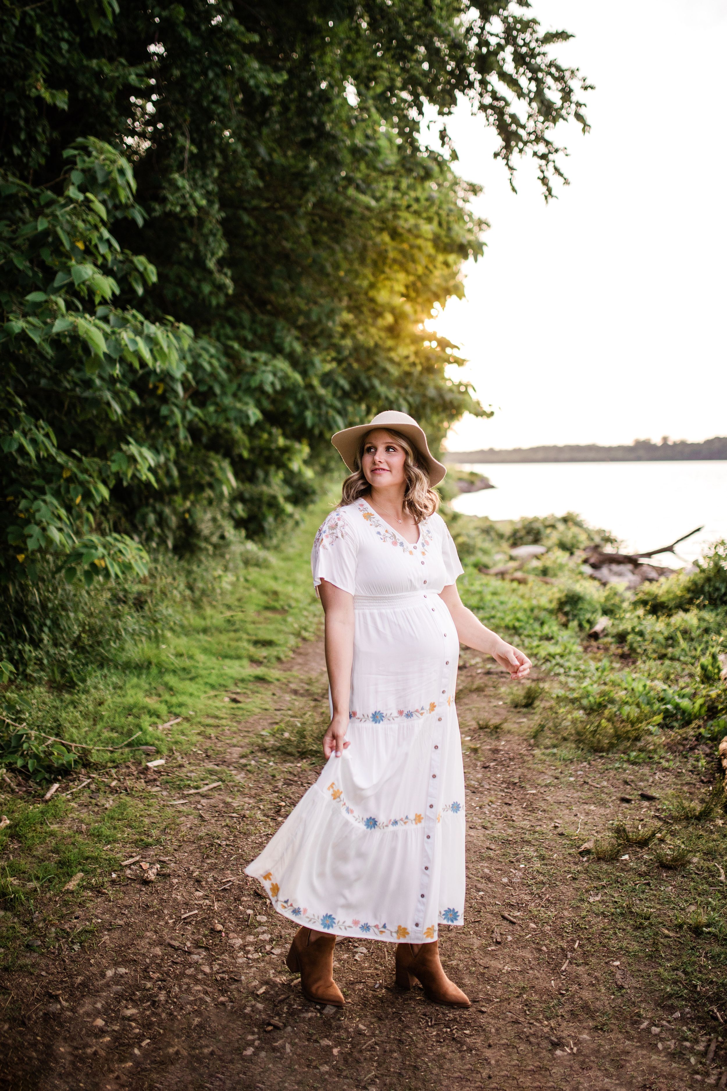 Pregnant woman in white embroidered maxi dress and straw hat walking along a lakeside trail.