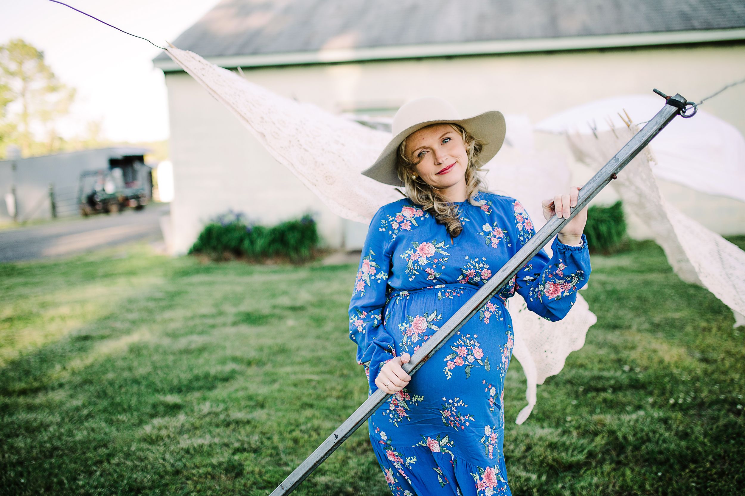 Smiling pregnant woman in blue floral dress holding clothesline pole outdoors.