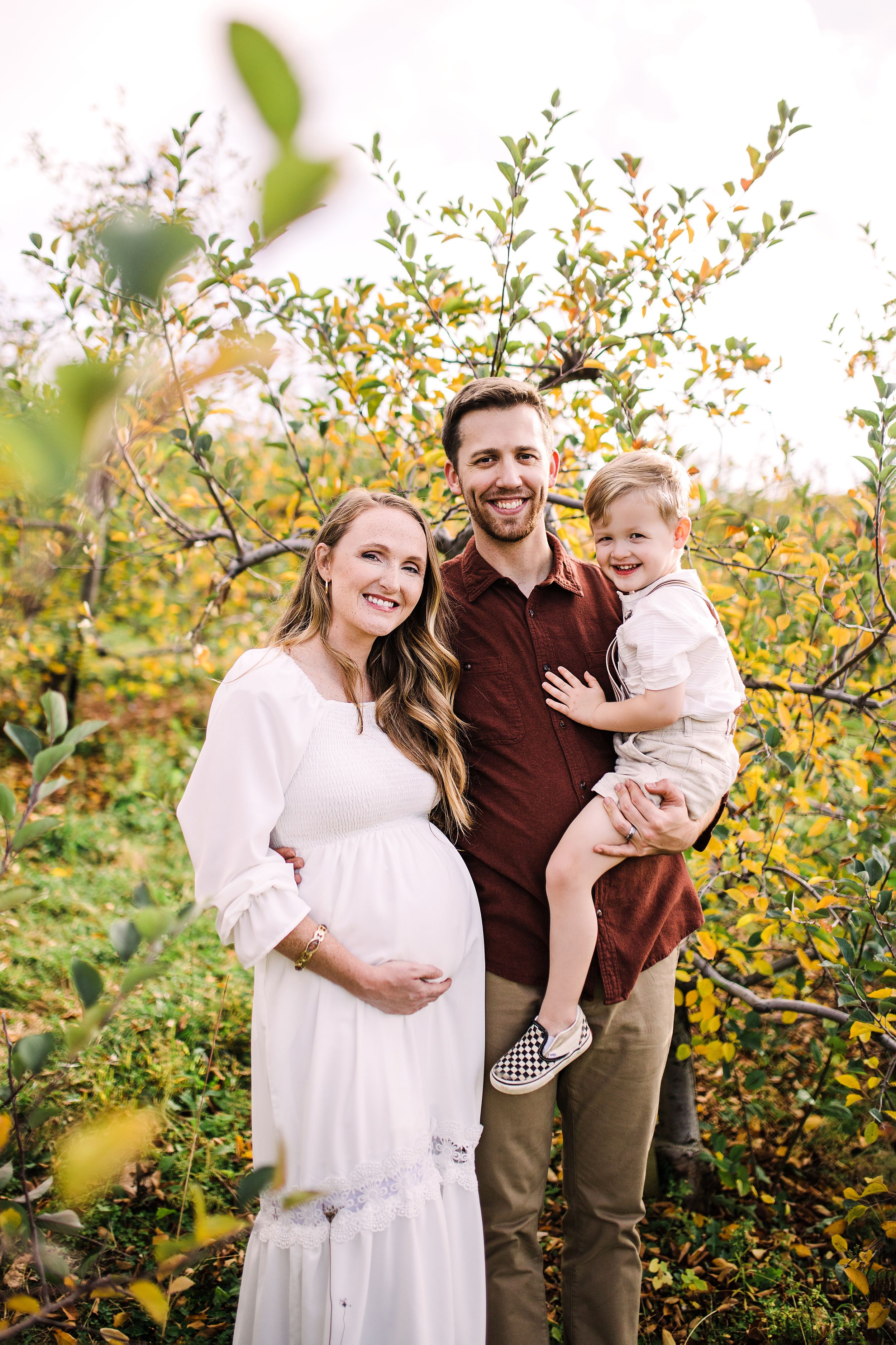Smiling pregnant woman in white dress with husband and toddler son in autumn orchard.