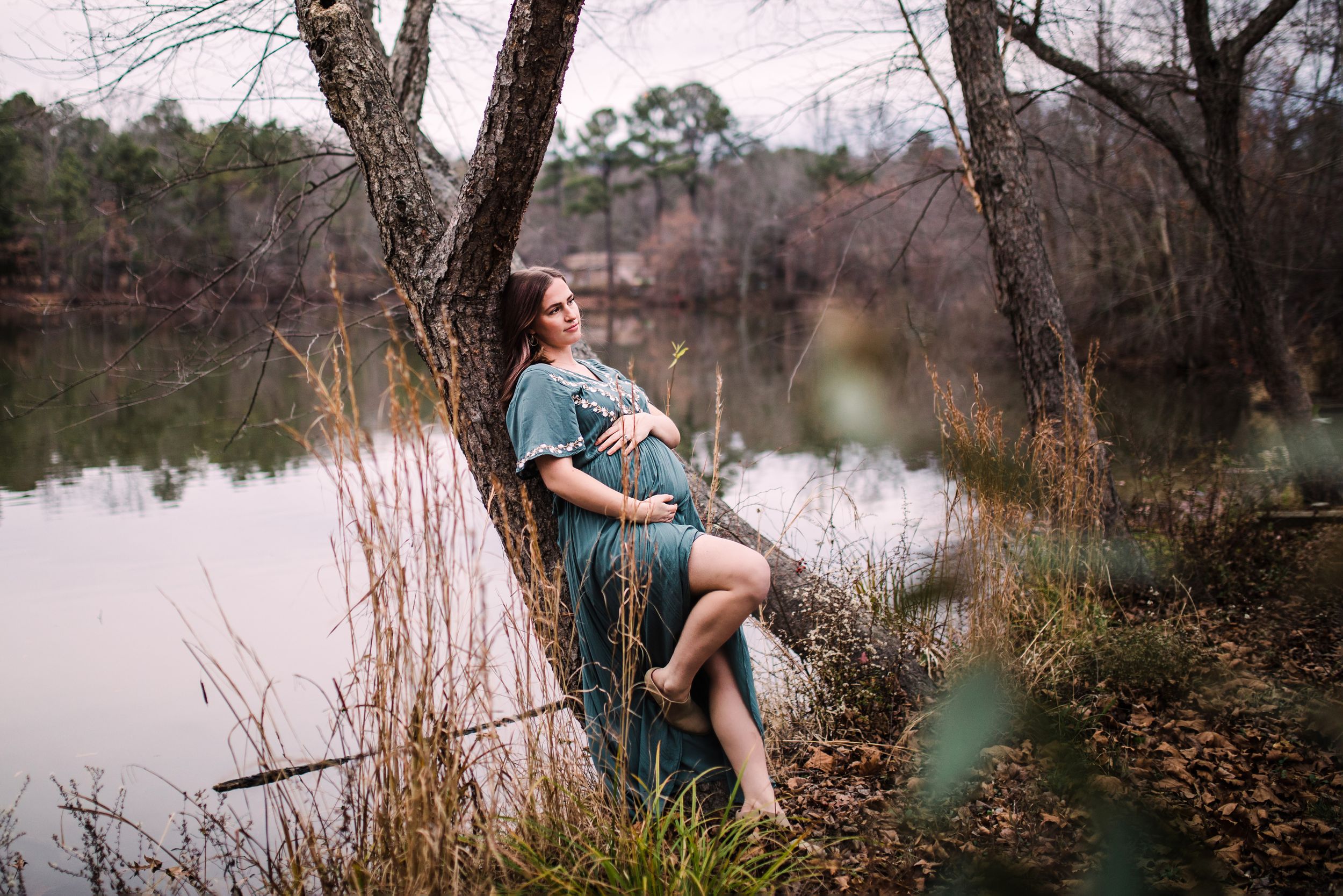 Barefoot pregnant woman in teal dress leaning against tree by tranquil forest lake.