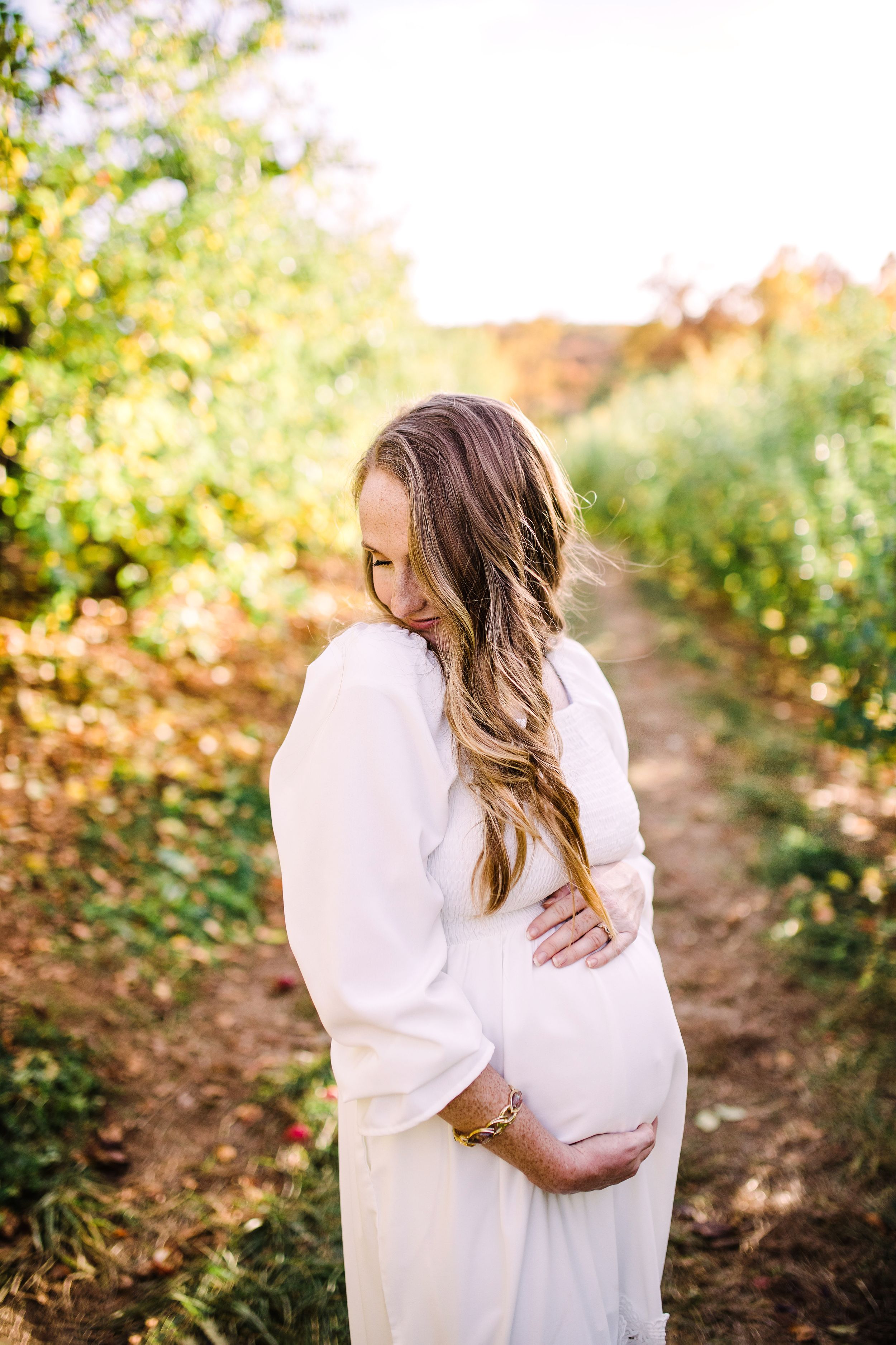 Pregnant woman in white dress cradling baby bump while looking down in sunlit orchard path.