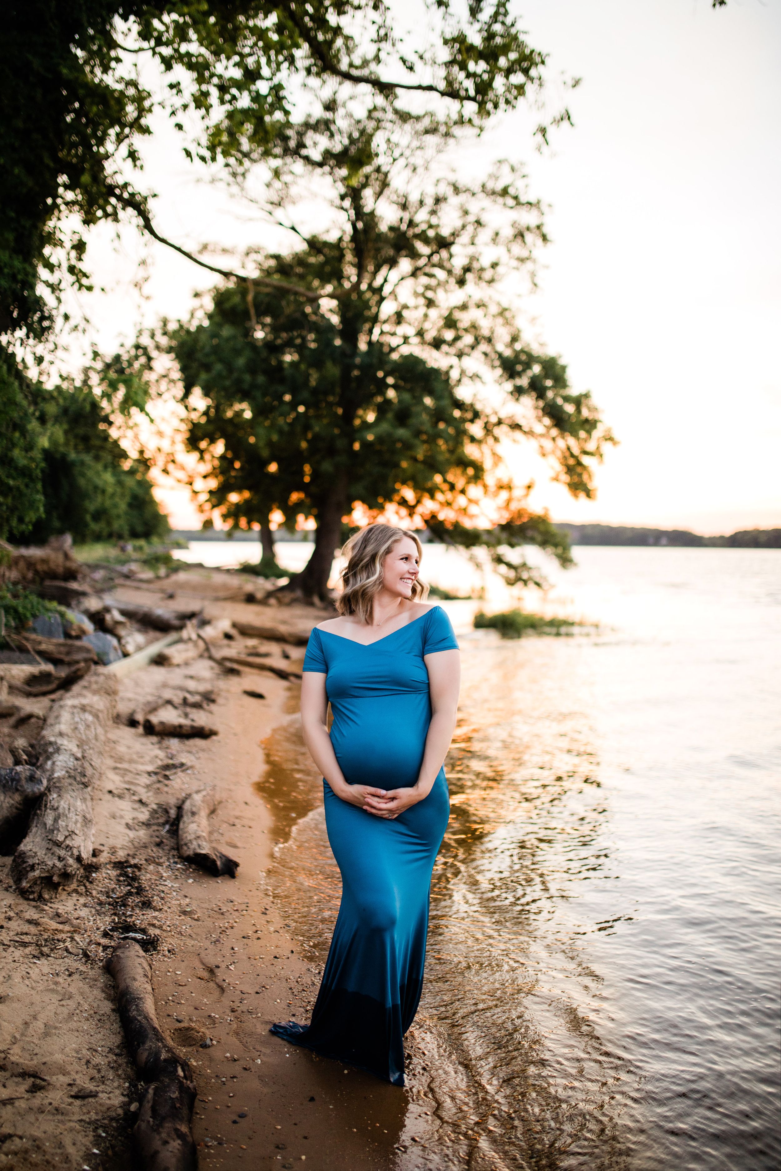 Pregnant woman in teal form-fitting gown standing on rocky lakeside shore at golden sunset.