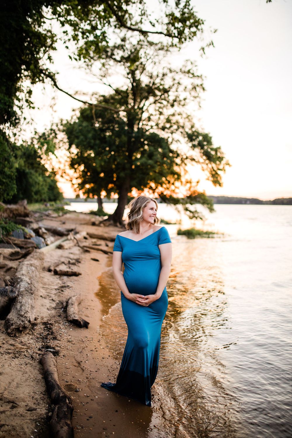 Pregnant woman in teal form-fitting gown standing on rocky lakeside shore at golden sunset.