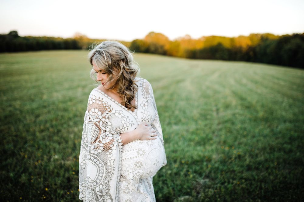 Pregnant woman in white crochet lace wrap gazing down at her bump in a green field at sunset.