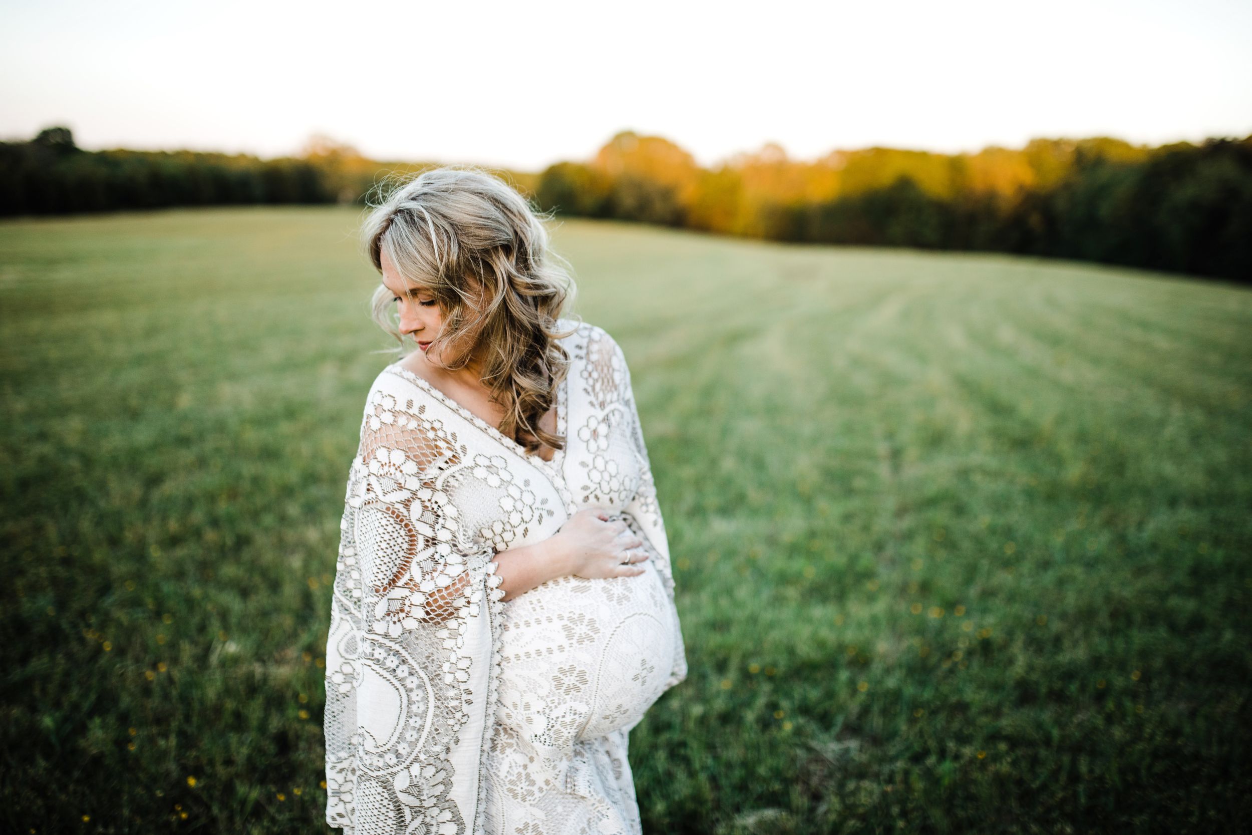 Pregnant woman in white crochet lace wrap gazing down at her bump in a green field at sunset.