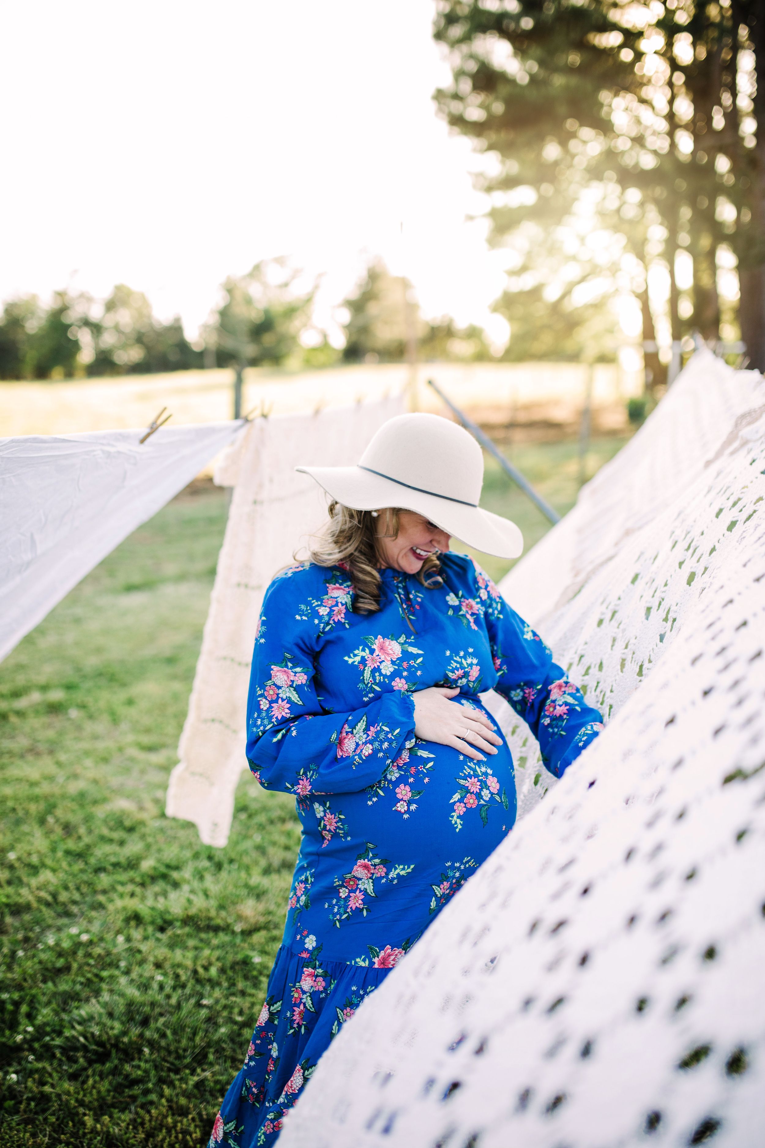 Smiling pregnant woman in blue floral dress and hat cradling baby bump.