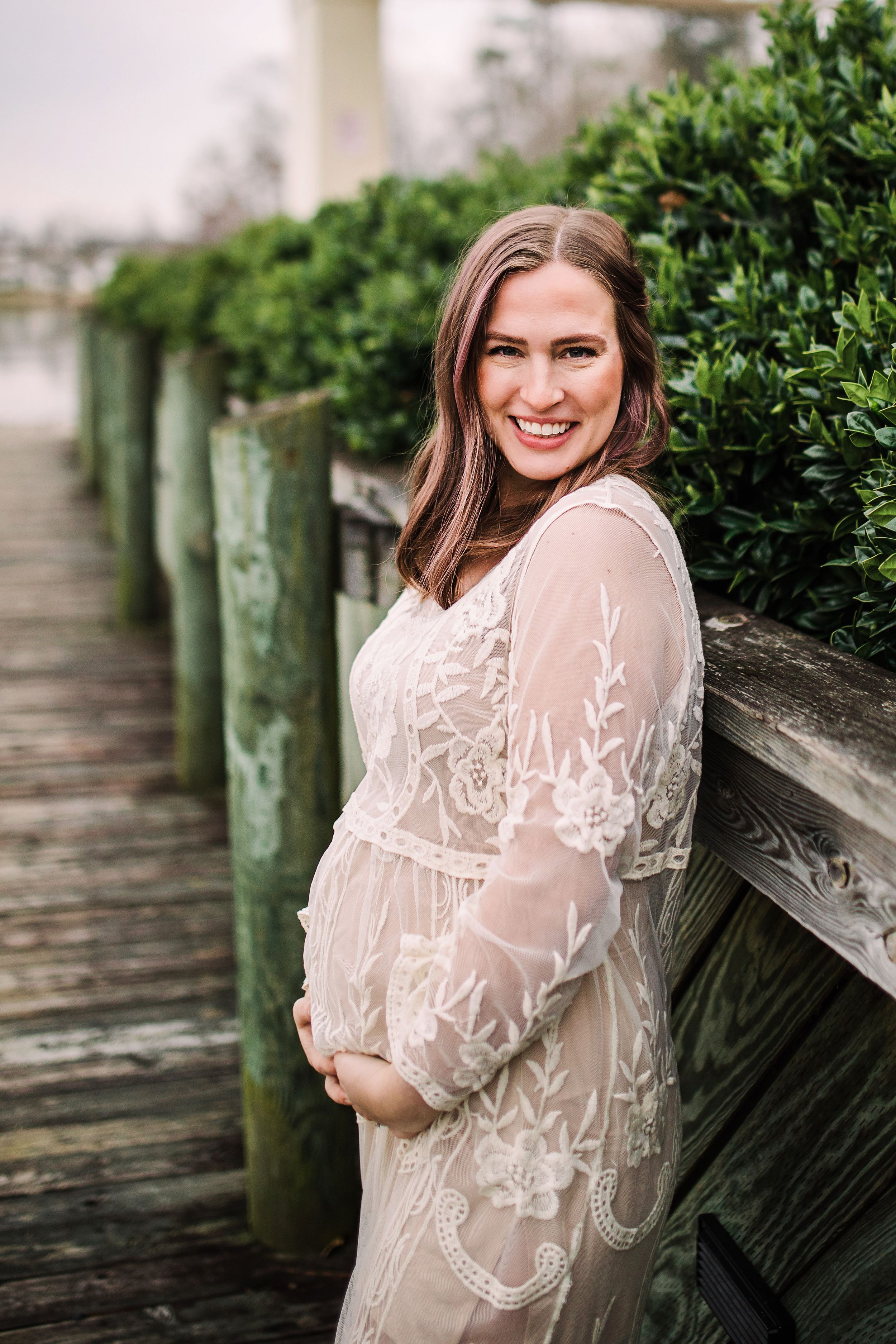 Smiling pregnant woman in white lace gown posing on a wooden dock surrounded by greenery.