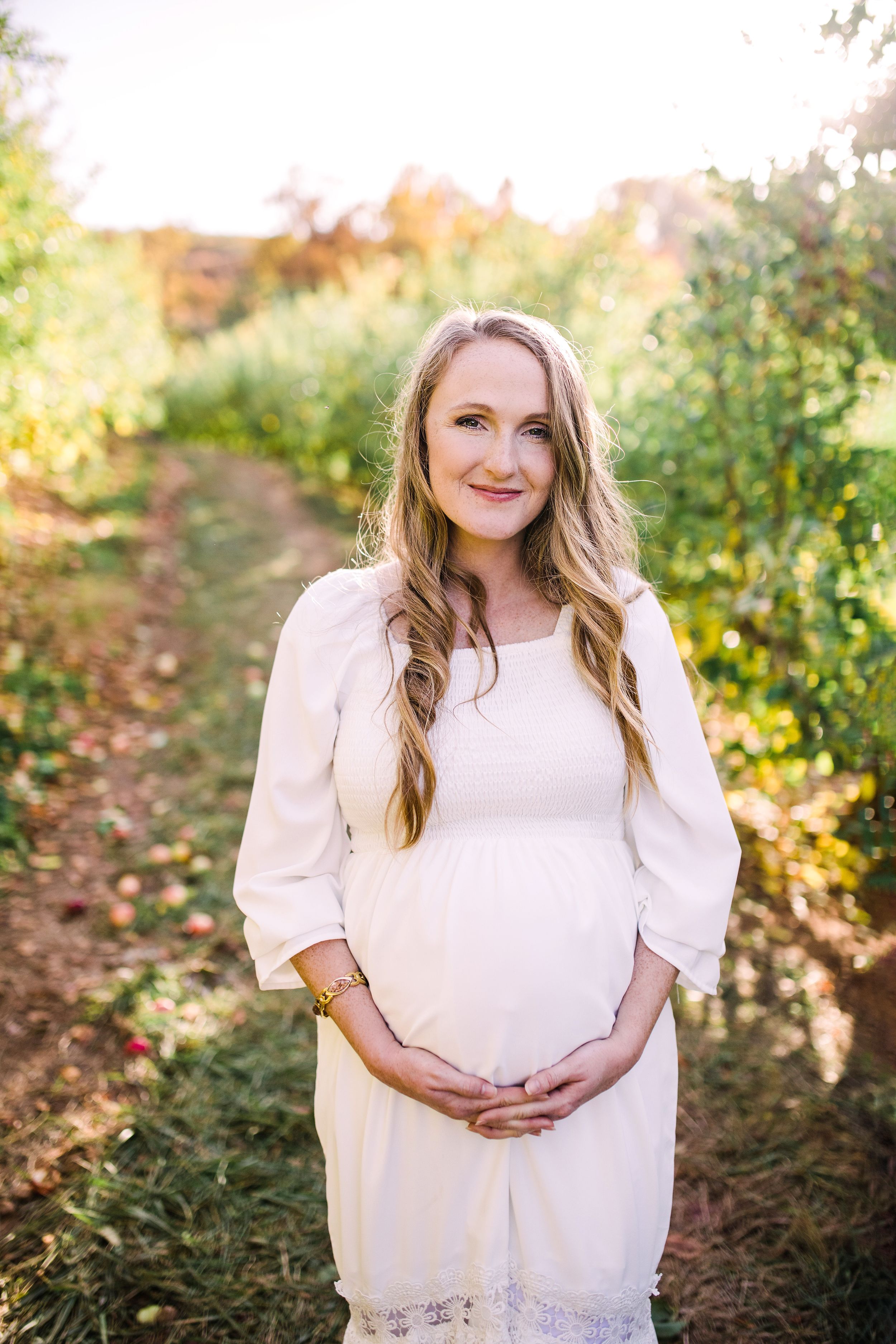 Smiling pregnant woman in white dress holding baby bump in warm sunlit apple orchard.