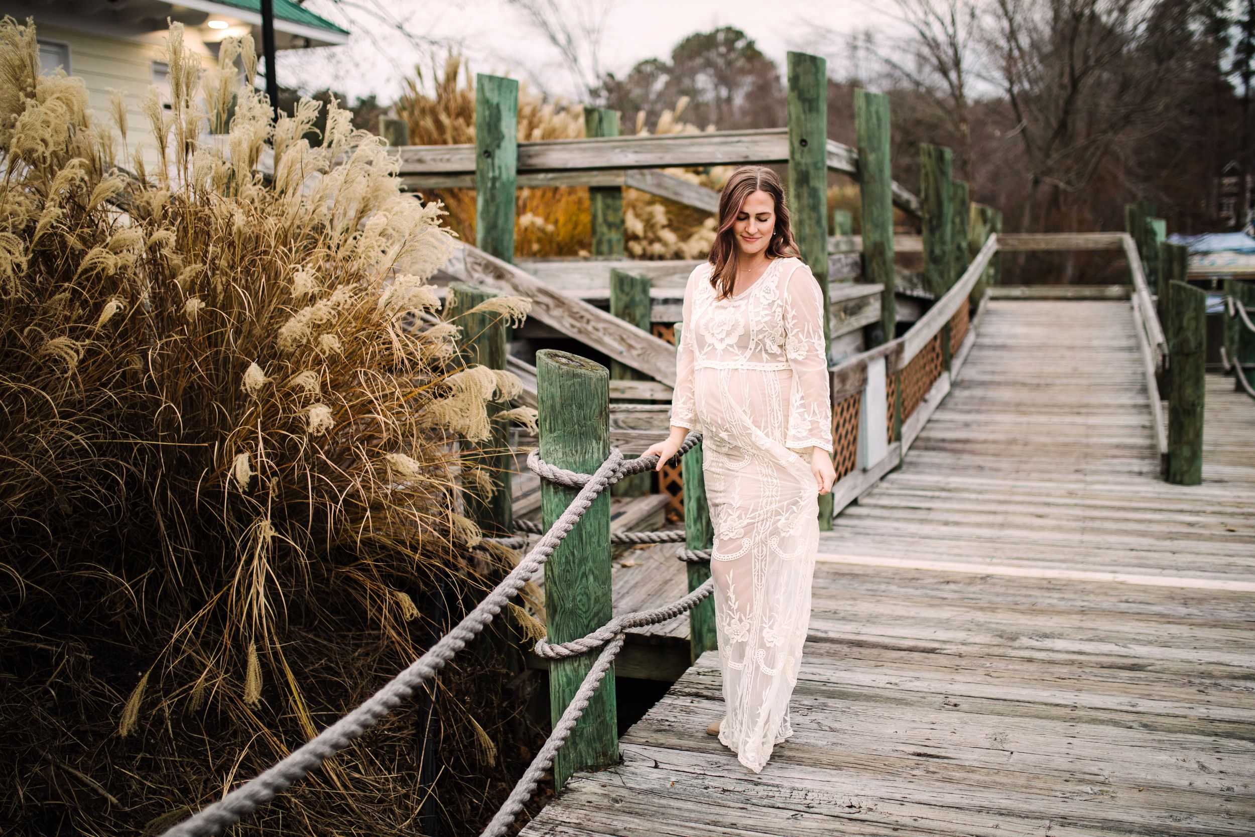 Pregnant woman in cream lace gown walking along rustic wooden dock surrounded by tall grasses.