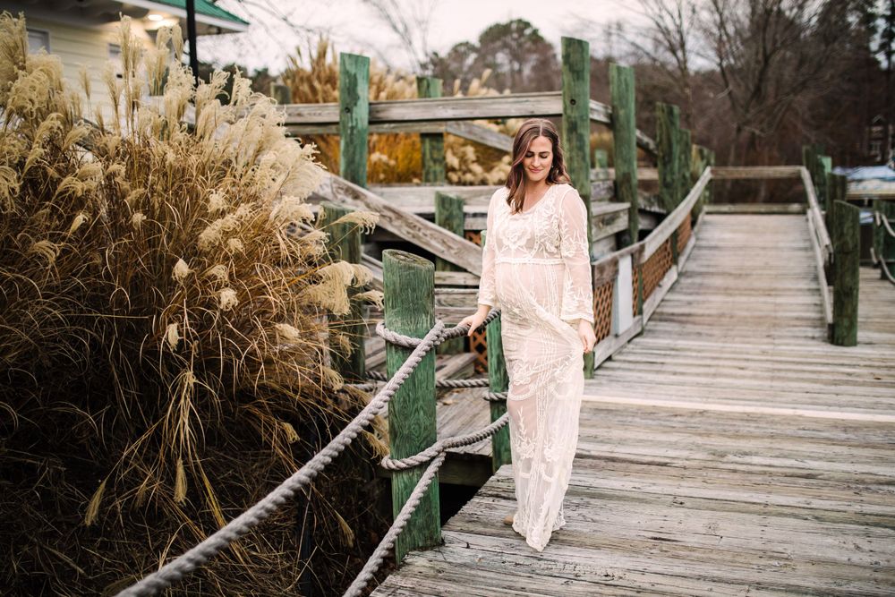 Pregnant woman in cream lace gown walking along rustic wooden dock surrounded by tall grasses.