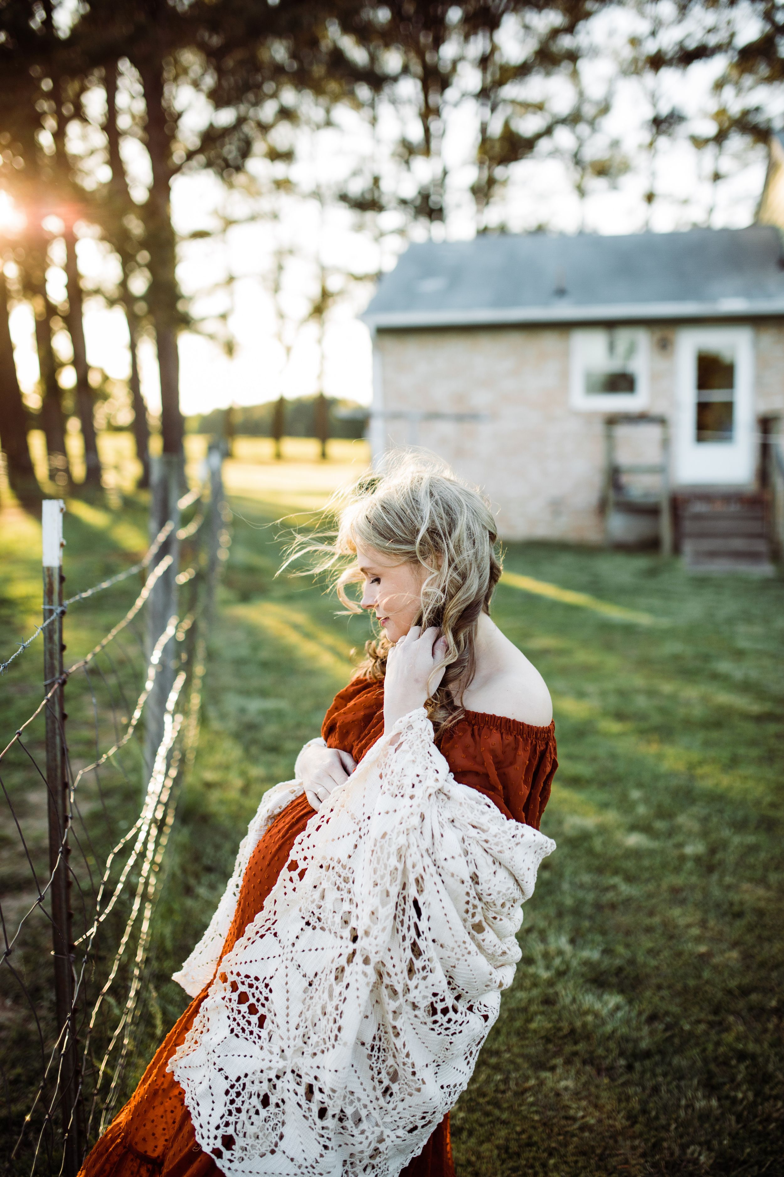Blonde pregnant woman in rust dress carrying white lace wrap near fence at golden sunset.