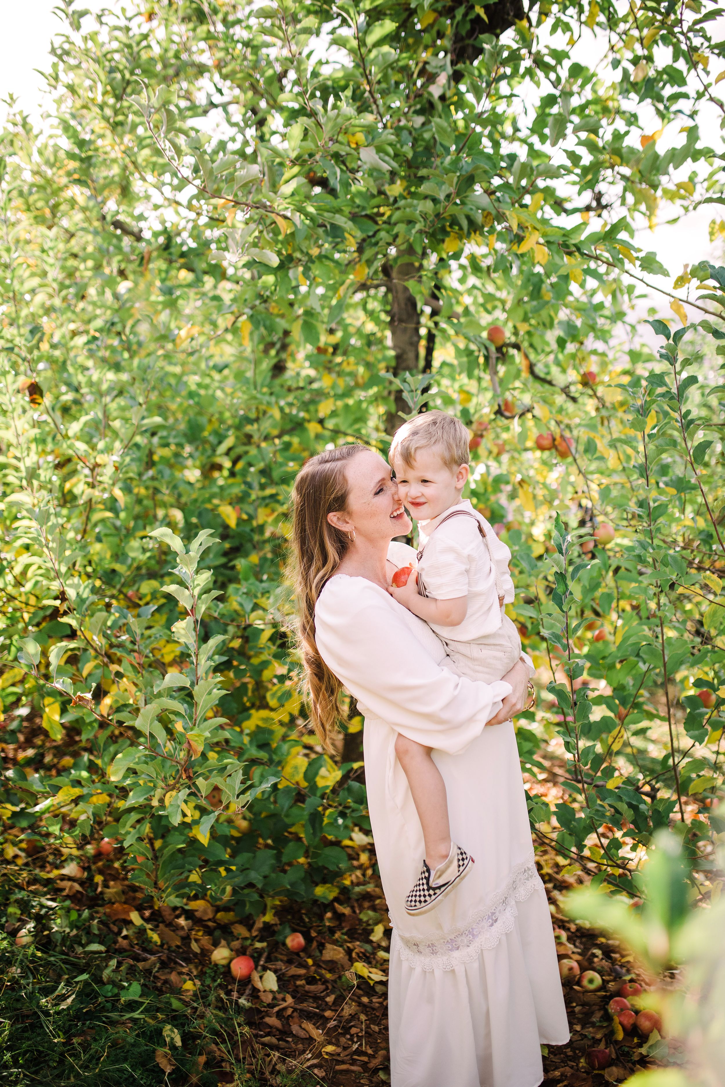 Pregnant woman in white dress lovingly holding laughing toddler boy in lush apple orchard.