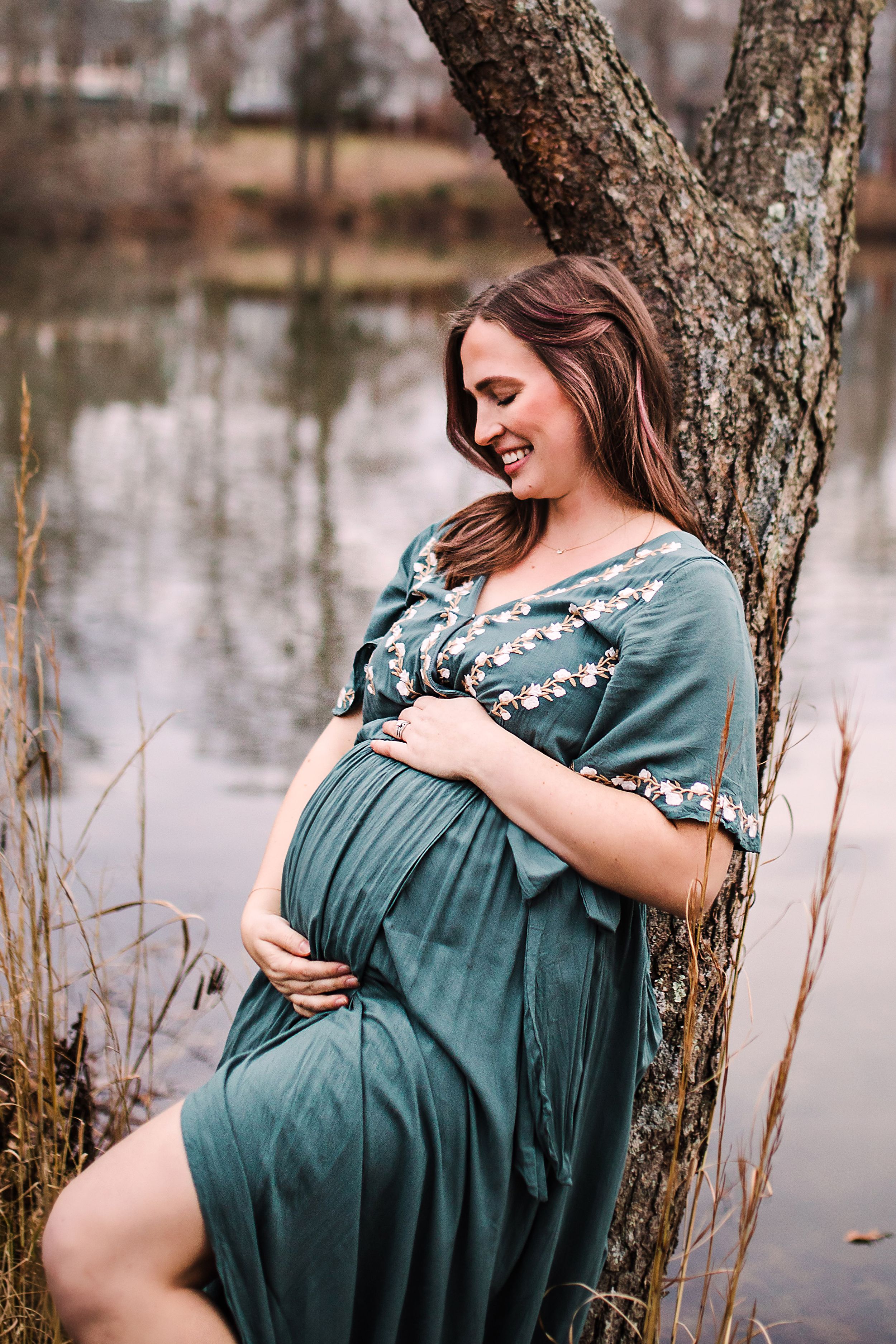 Smiling pregnant woman in teal embroidered dress cradling baby bump while leaning against tree by serene lake.
