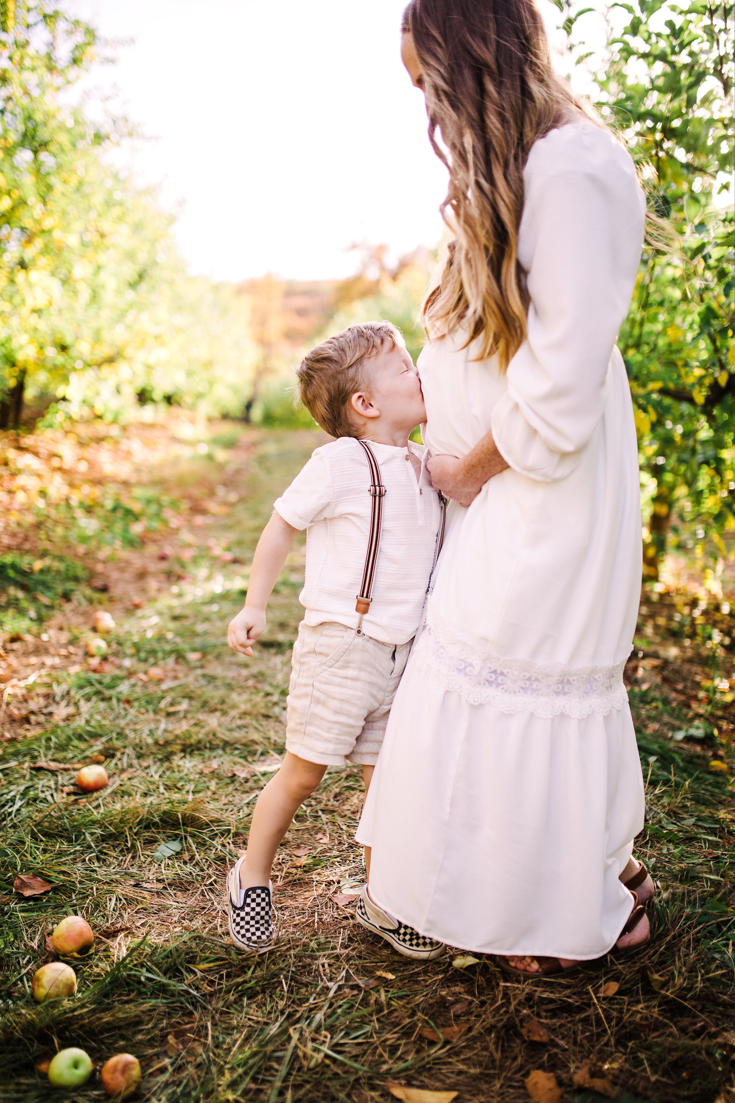 Young boy kissing pregnant mothers belly outdoors in sunny apple orchard during maternity session.