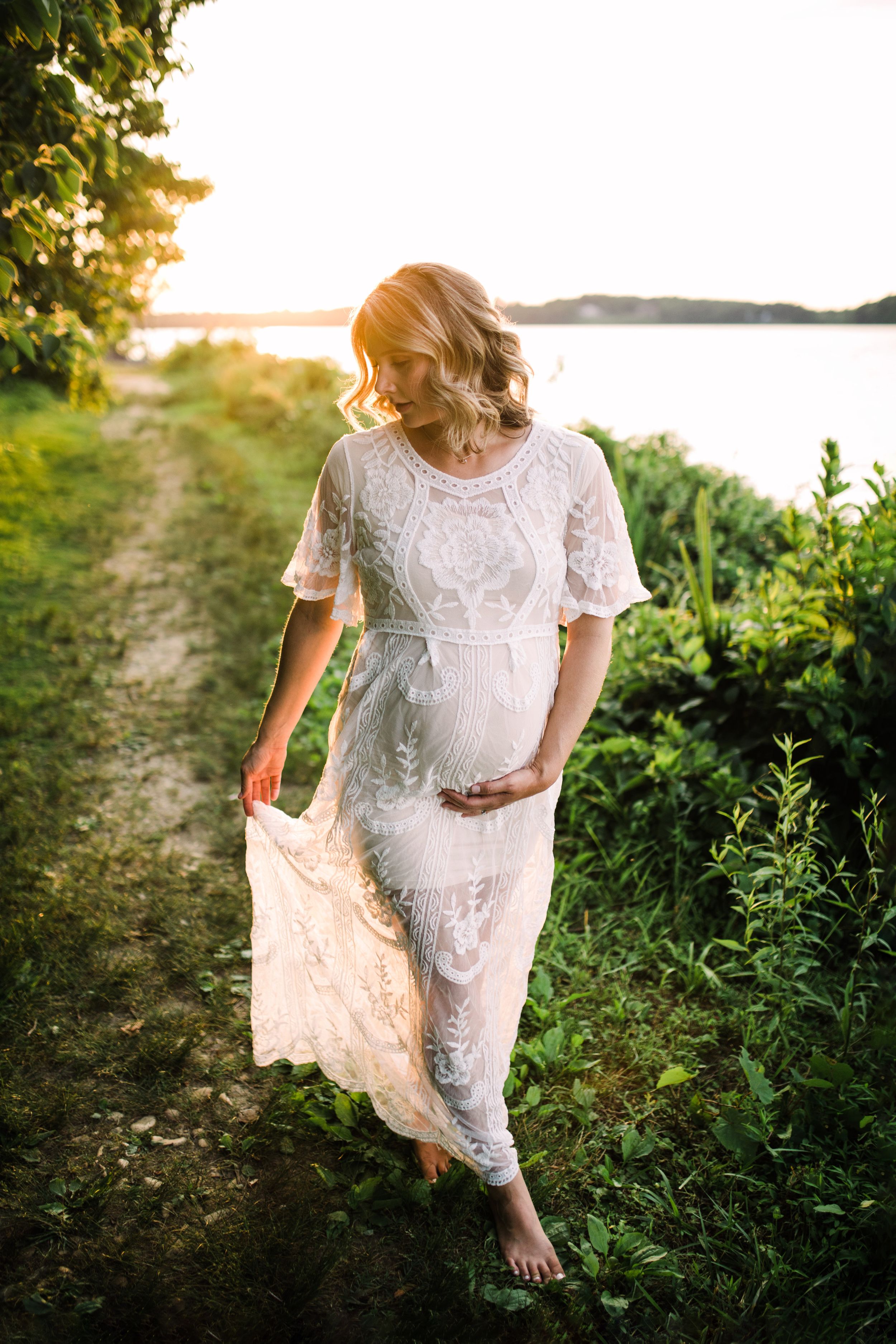 Pregnant woman in white lace dress walking barefoot along a sunlit lakeside path at sunset.