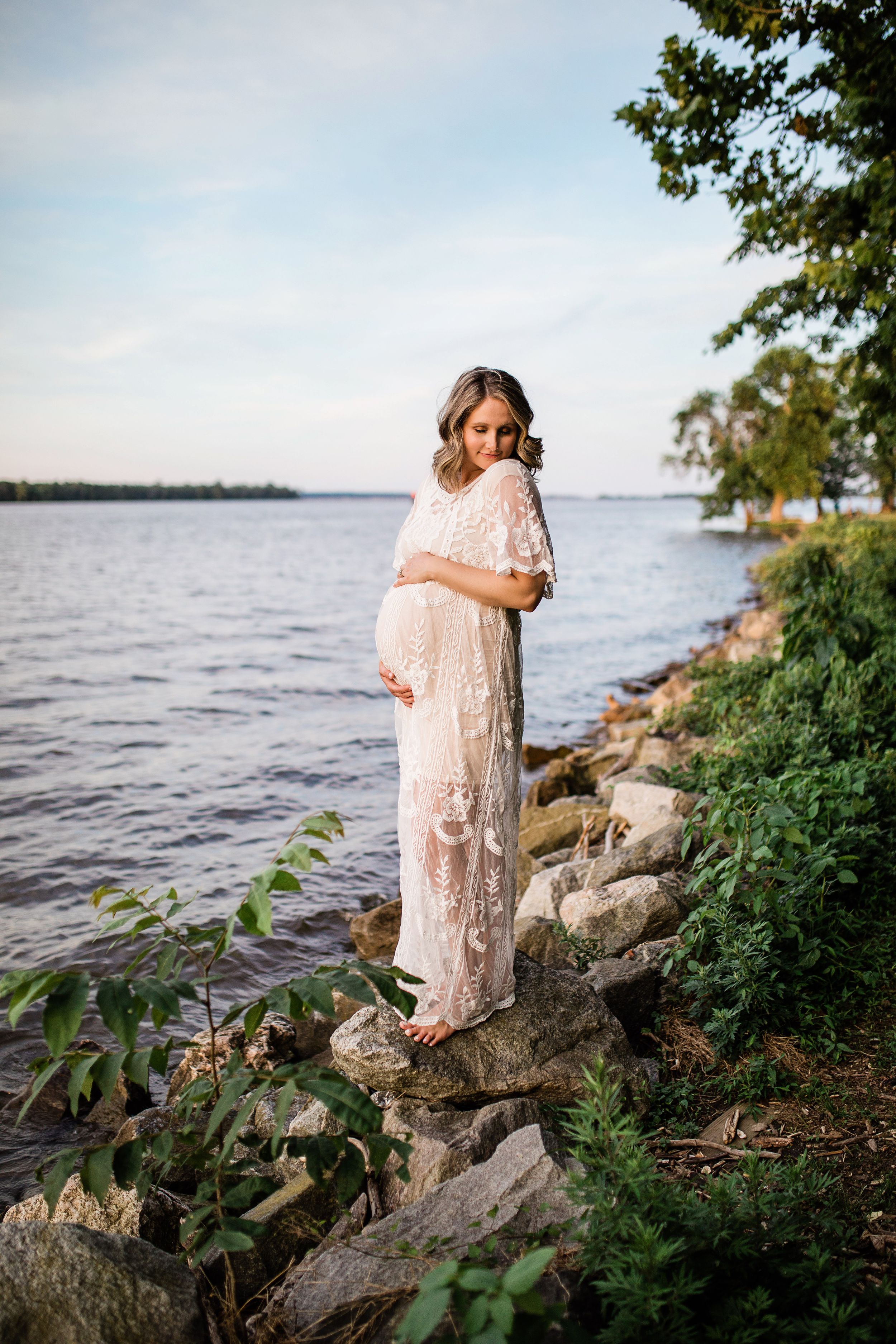 Pregnant woman in white lace dress standing on rocky shoreline by a calm lake at dusk.