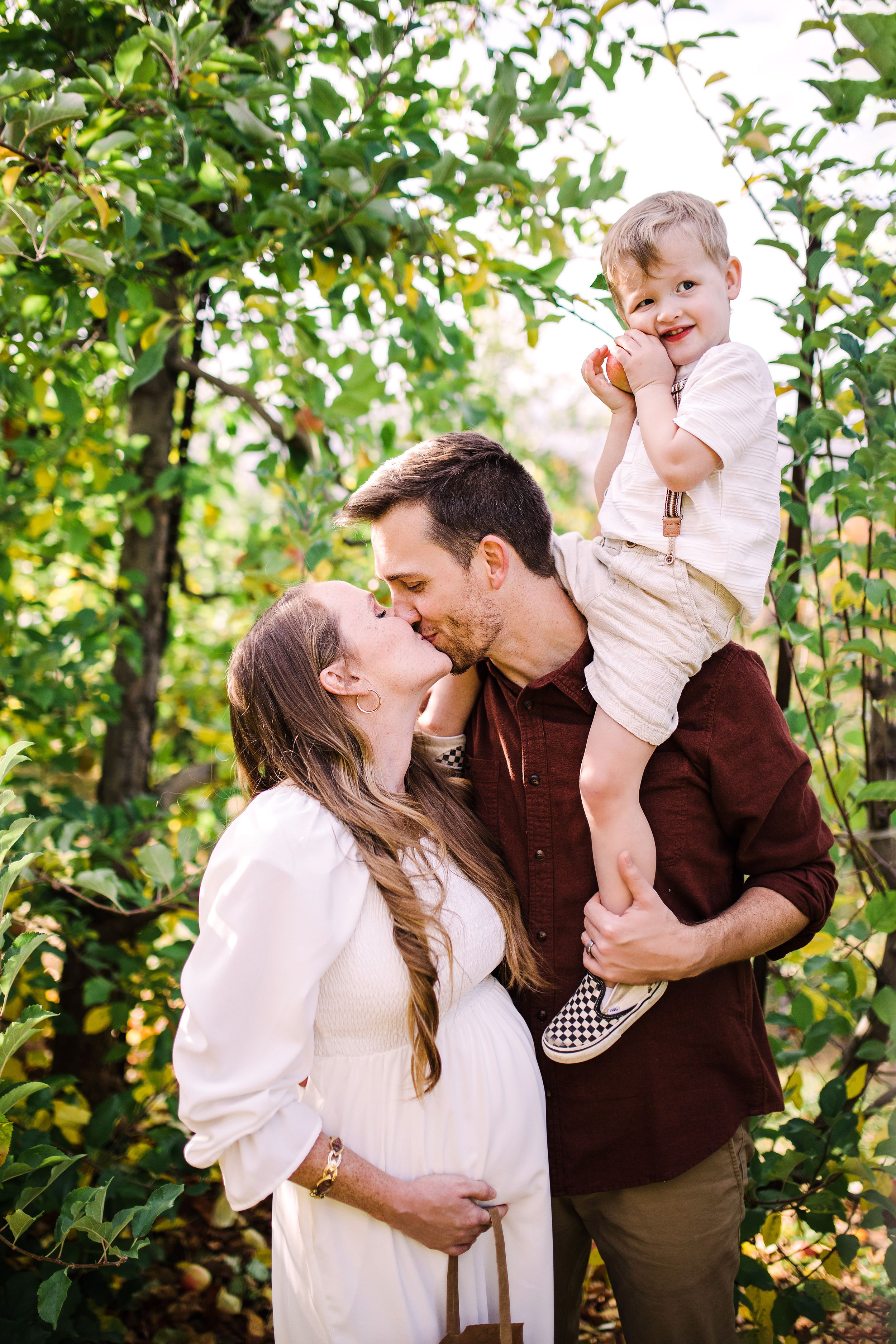 Couple kissing while man holds toddler son during outdoor maternity photo session in green orchard.