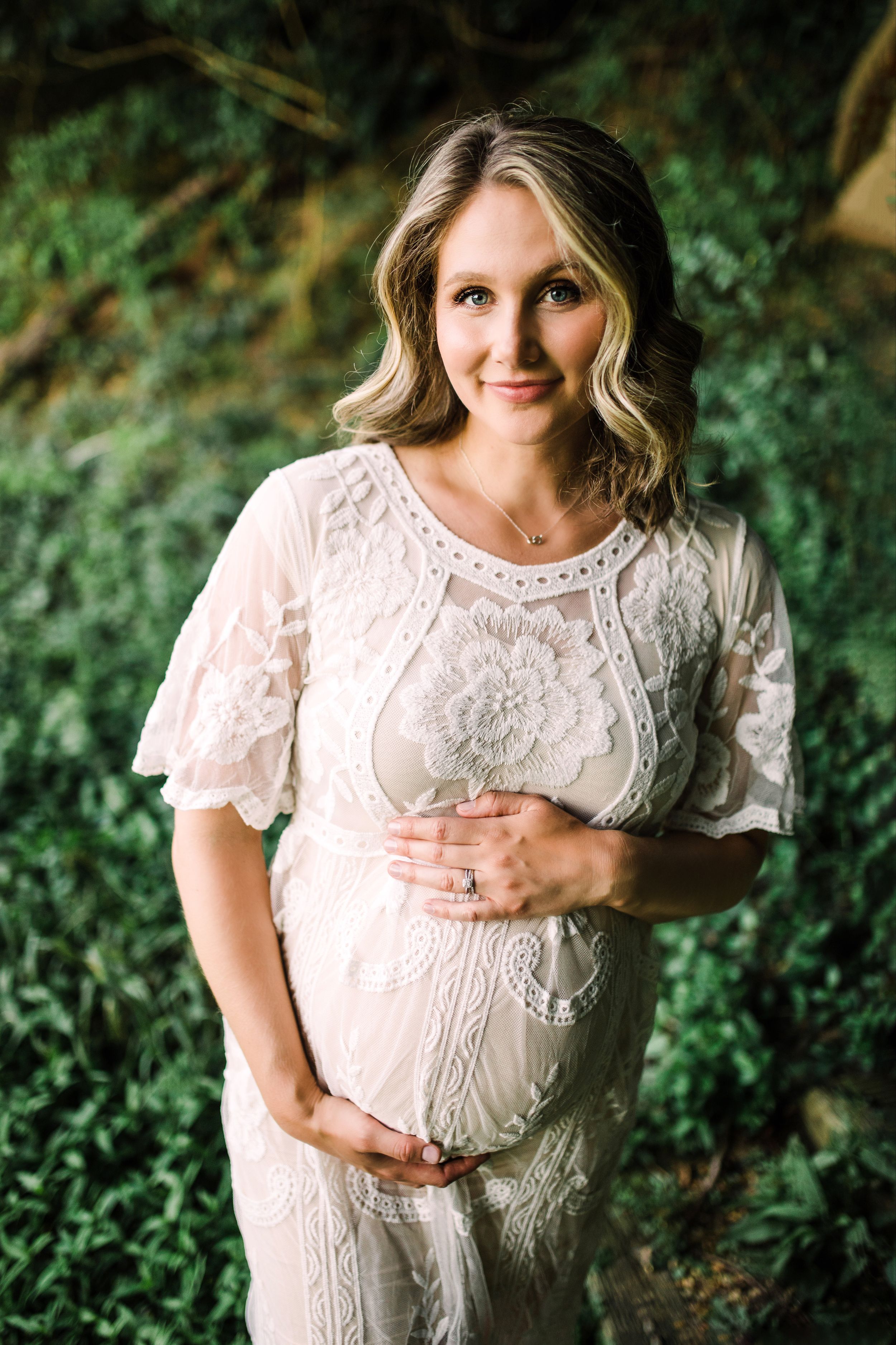 Smiling pregnant woman in white lace flutter-sleeve dress standing before lush green foliage.