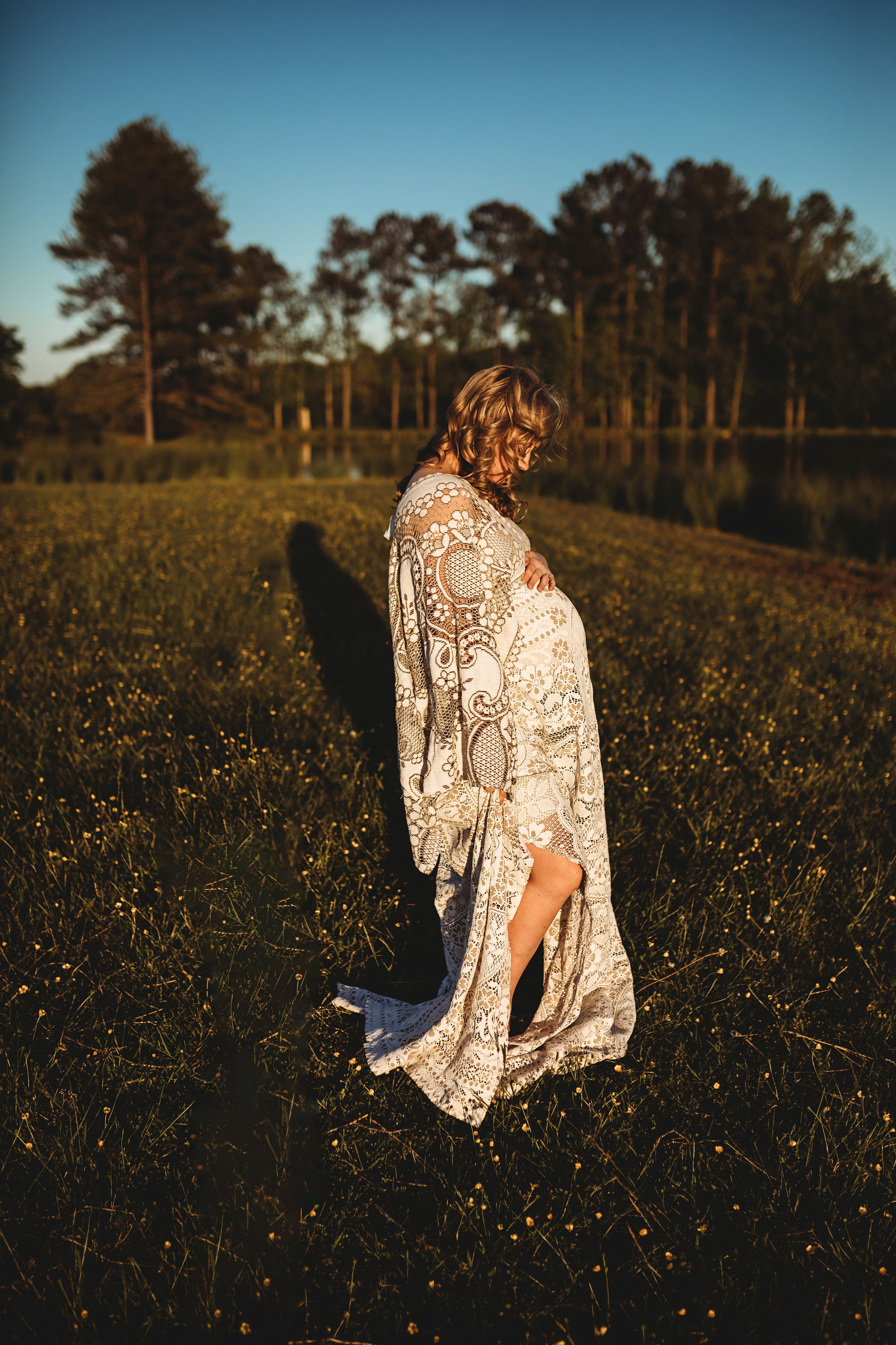Pregnant woman in bohemian lace robe standing in a sunlit wildflower field near pine trees.