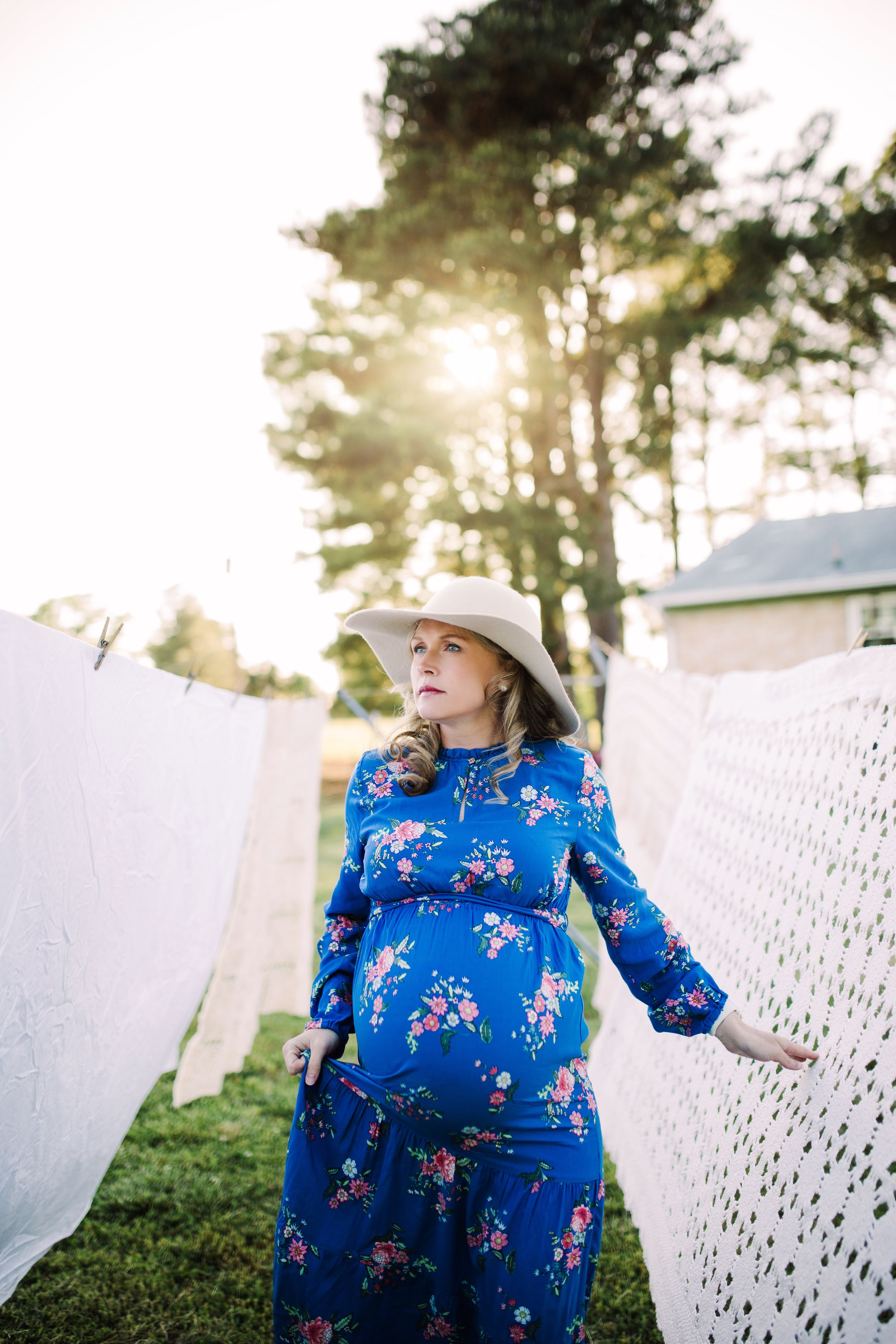 Pregnant woman in blue floral dress and hat by clothesline at sunset.