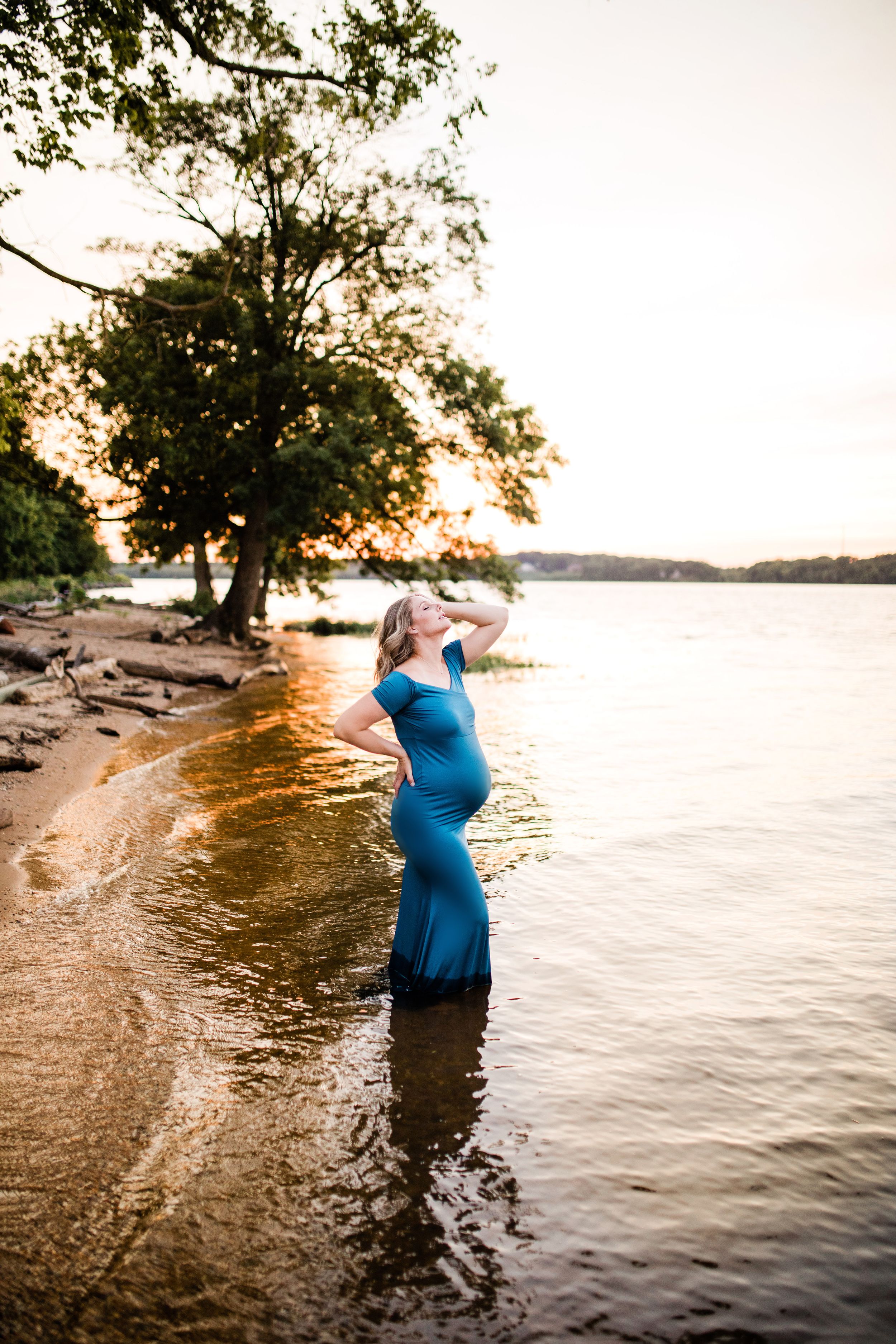 Pregnant woman in teal dress standing in lake water at golden sunset.