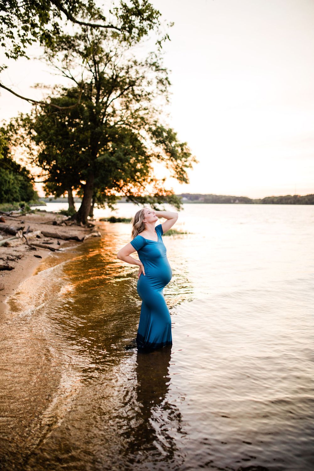 Pregnant woman in teal dress standing in lake water at golden sunset.