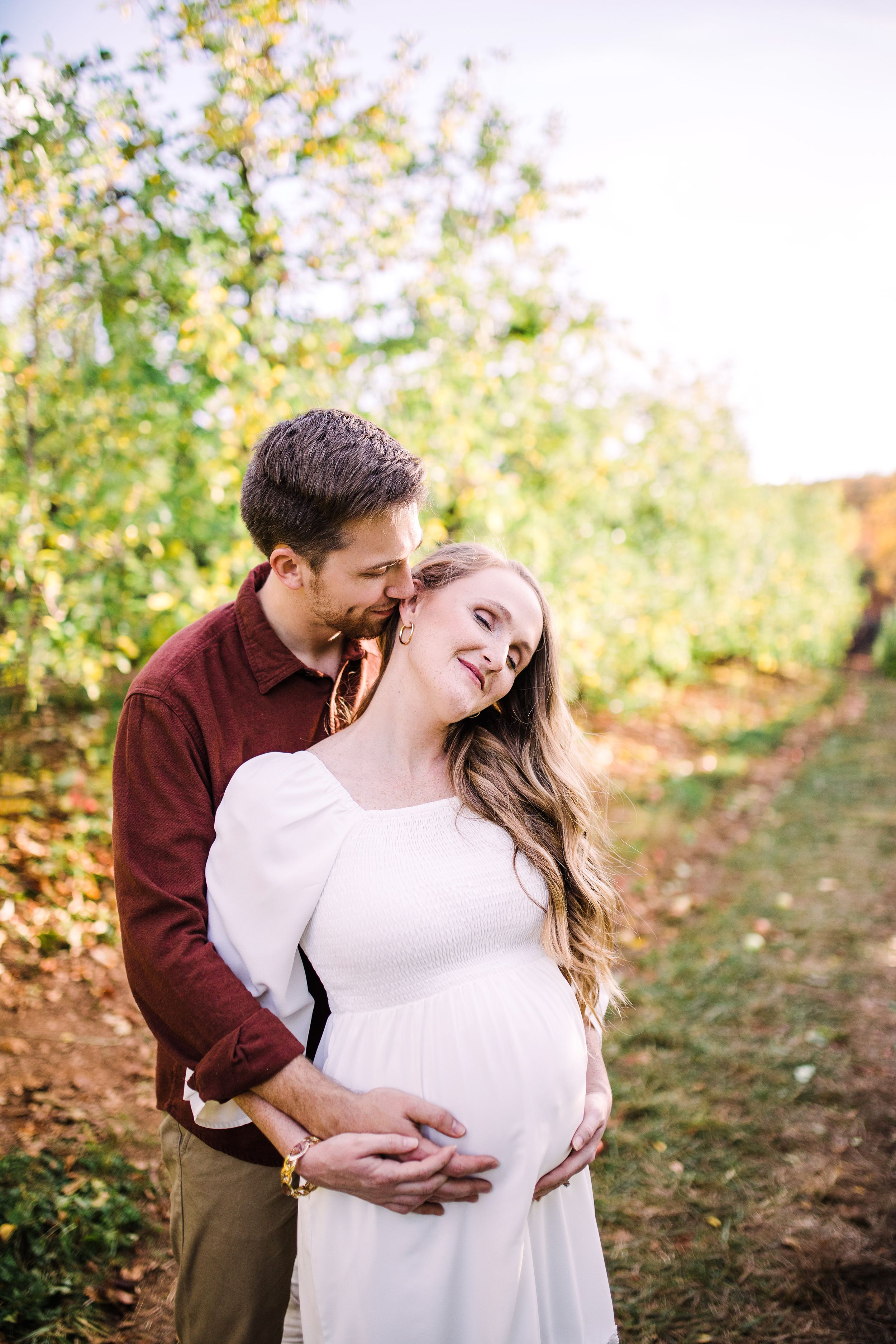 Man tenderly embracing pregnant wife from behind in golden sunlit orchard maternity portrait.